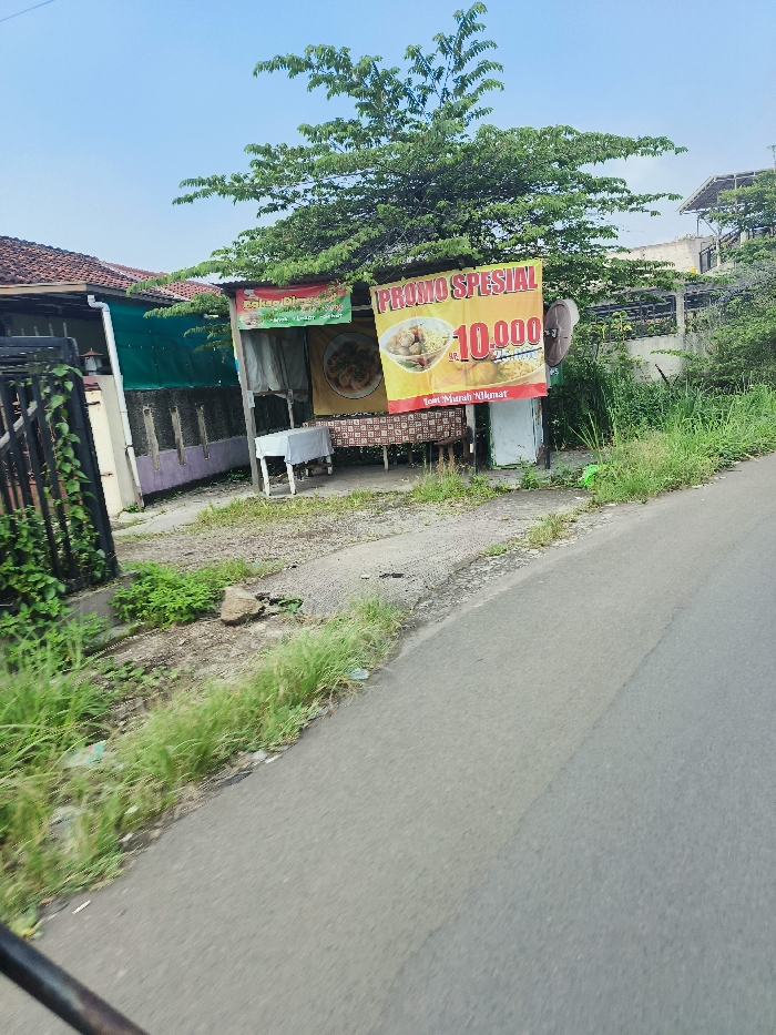 The image shows a street-side food stall in Cileunyi, Indonesia, located at coordinates -6.946 latitude and 107.725 longitude. The stall is a small, open-air structure with a yellow sign that reads "PROMO SPESIAL 10.000." The sign is adorned with an image of a bowl of food, likely meatballs. The stall is situated next to a small, single-story house with a green roof. The house is surrounded by lush vegetation, and a black metal fence separates the property from the street. In the foreground, a paved road extends from the bottom left of the frame toward the right. There are no people visible in the image. The sky is blue and cloudless.