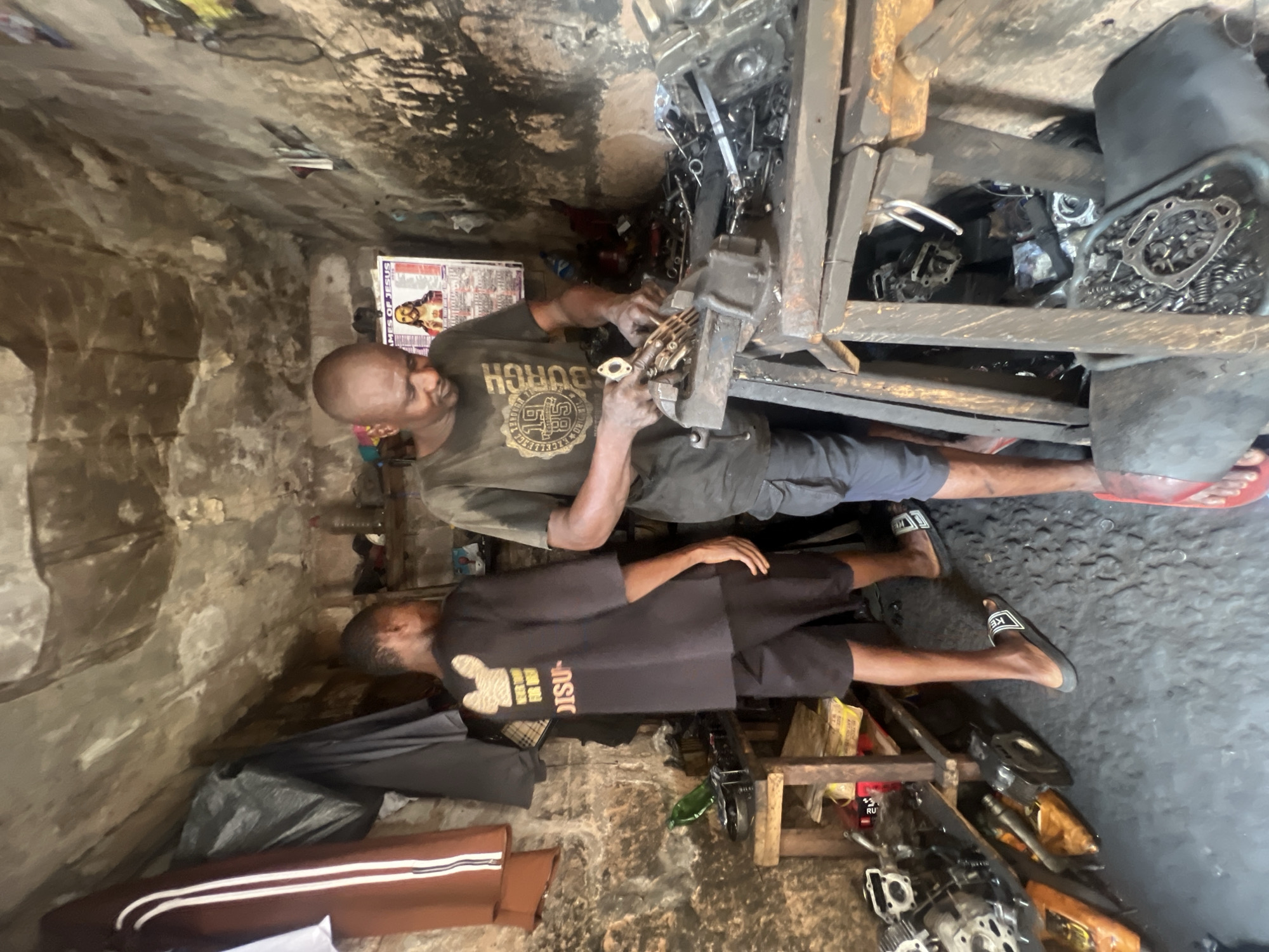 The image depicts two adult Black men in a cluttered, poorly lit workshop in Onitsha, Nigeria. The older man, wearing a dark t-shirt with gold lettering, is working on a motorcycle engine clamped in a metal vise. Parts and tools surround him. The younger man, wearing a dark t-shirt with gold lettering and shorts, lies on a makeshift wooden bench near the first man. The workshop has bare concrete walls, and the floor is stained with oil. Various motorcycle parts and tools are scattered around the room. A poster featuring a religious image is affixed to the wall behind the older man. A plastic bag sits near the younger man's feet.