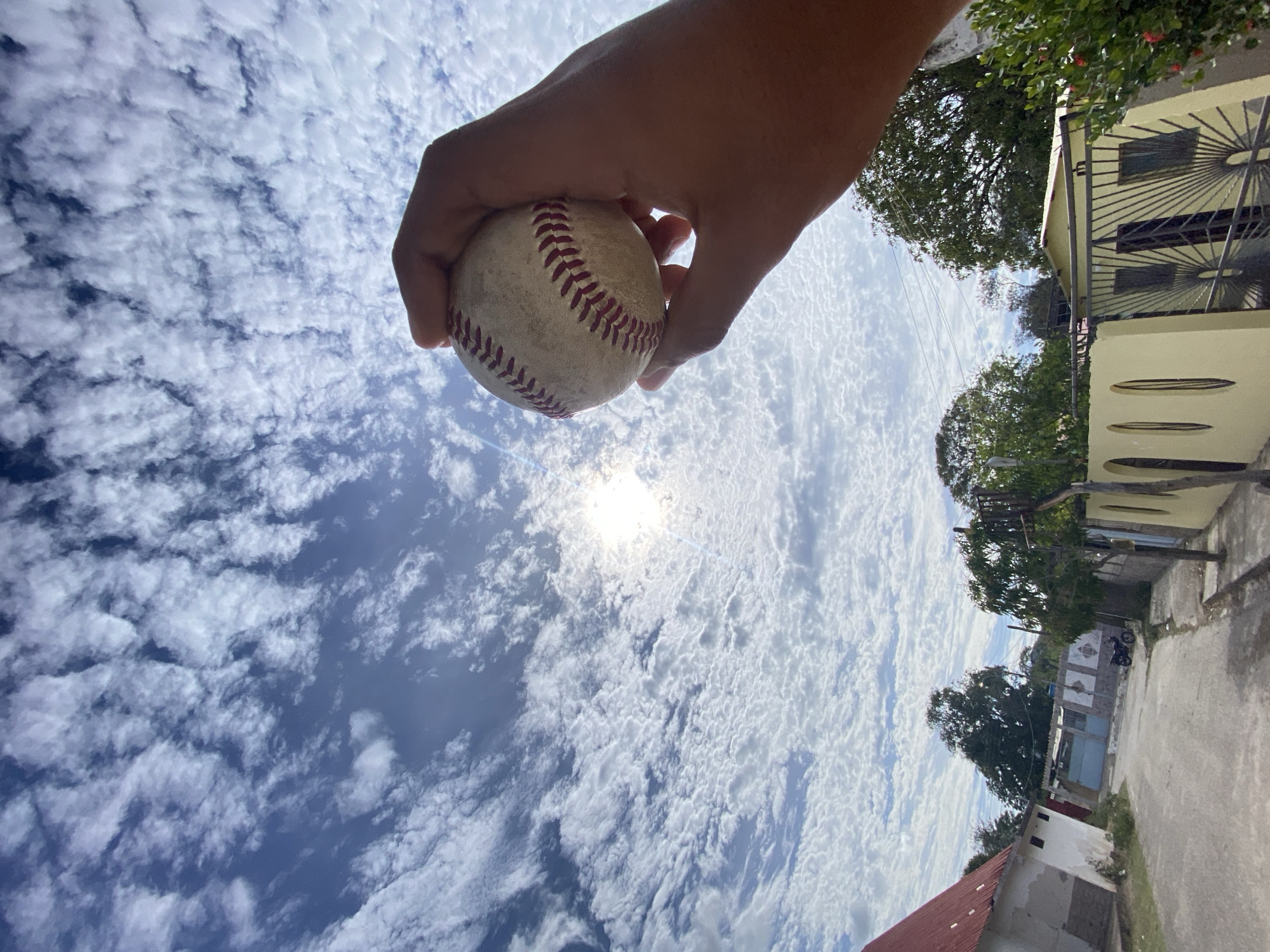 A low-angle outdoor shot depicts a baseball held in a hand against a partly cloudy sky. The sun is shining through the clouds, creating a bright spot near the center. The hand is holding the baseball between the thumb and fingers. In the background, blurred buildings and trees suggest a residential area. The image is rotated, showing elements of a city street or alley. The location is Juan Griego, Venezuela, as implied by the prompt. The angle of the shot obscures details about the people. There's an asphalt street and some building architecture typical of a low-income suburban location, including a concrete curb.