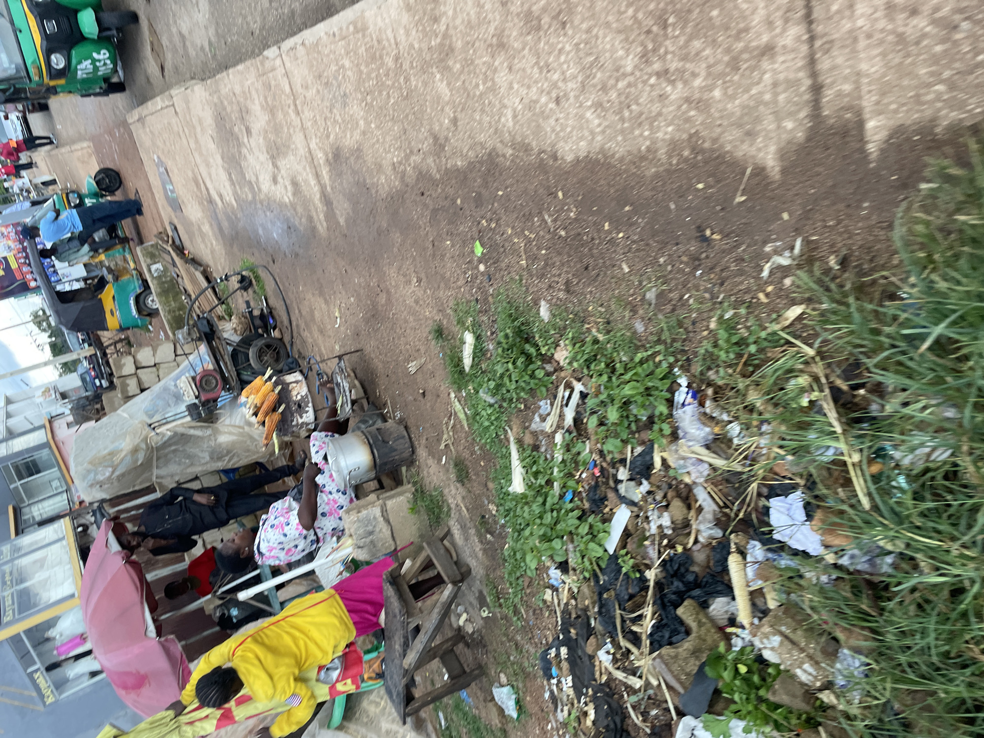 The image depicts a street scene in Jos, Nigeria, characterized by a roadside vendor and surrounding urban environment. In the foreground, a pile of refuse, including plastic bags, wrappers, and organic debris, is visible alongside overgrown grass.
Central to the scene is a food stall with a pink umbrella. A woman wearing a floral dress is seated, and a man in a black outfit stands nearby, appearing to engage in conversation. In front of them, ears of corn are being roasted over a fire. To the left, a person in a yellow jacket and pink skirt stands near a small wooden stool.
Further down the street, three-wheeled auto-rickshaws, commonly known as "Keke Napep," are parked. One is green and yellow, with license plate "PTA 1156." Another Keke is visible behind it, also green and yellow. Several other individuals are present, some standing near the Keke, suggesting a public transportation hub or a busy thoroughfare. The background shows buildings and more Keke Napeps, indicating a commercial or densely populated area of the city. The overall impression is of a typical, lively street scene in Jos, with the presence of street food vendors and local transportation.