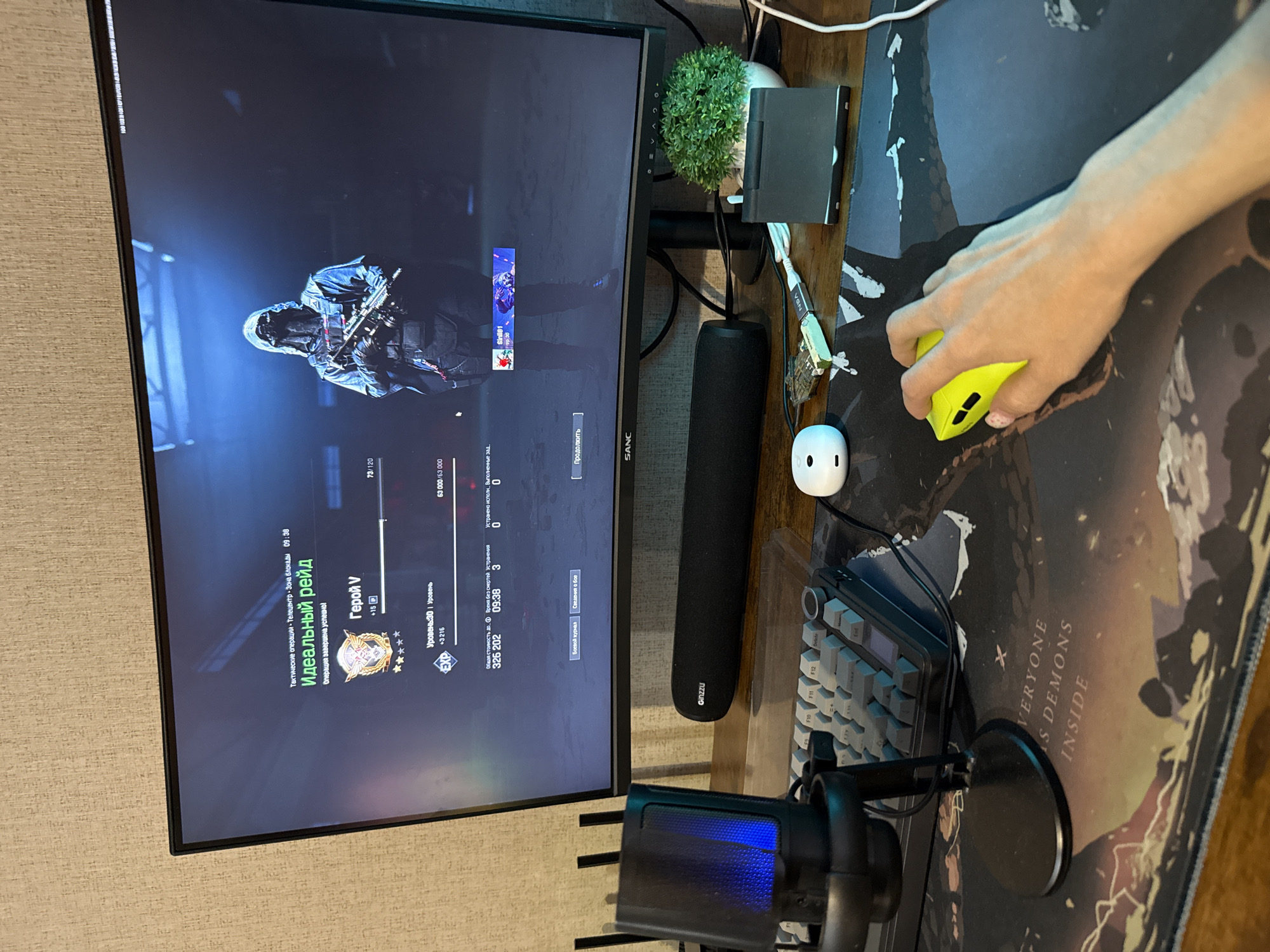A person's hand is gripping a yellow mouse on a desk, which is covered by a decorative mouse pad with the phrase "EVERYONE HAS DEMONS INSIDE". A computer monitor shows gameplay of a first-person shooter set in an urban environment, possibly Rusanovo, Russia, indicated by the Cyrillic text on the screen and visible objects. The game displays statistics like "Perfect Raid", "Hero V", level 30, and various in-game metrics.
The desk also holds a keyboard, microphone, small plant, a white USB hub, and a black wrist rest branded "Ginzzu". Cables are visible, connecting the peripherals to the computer.