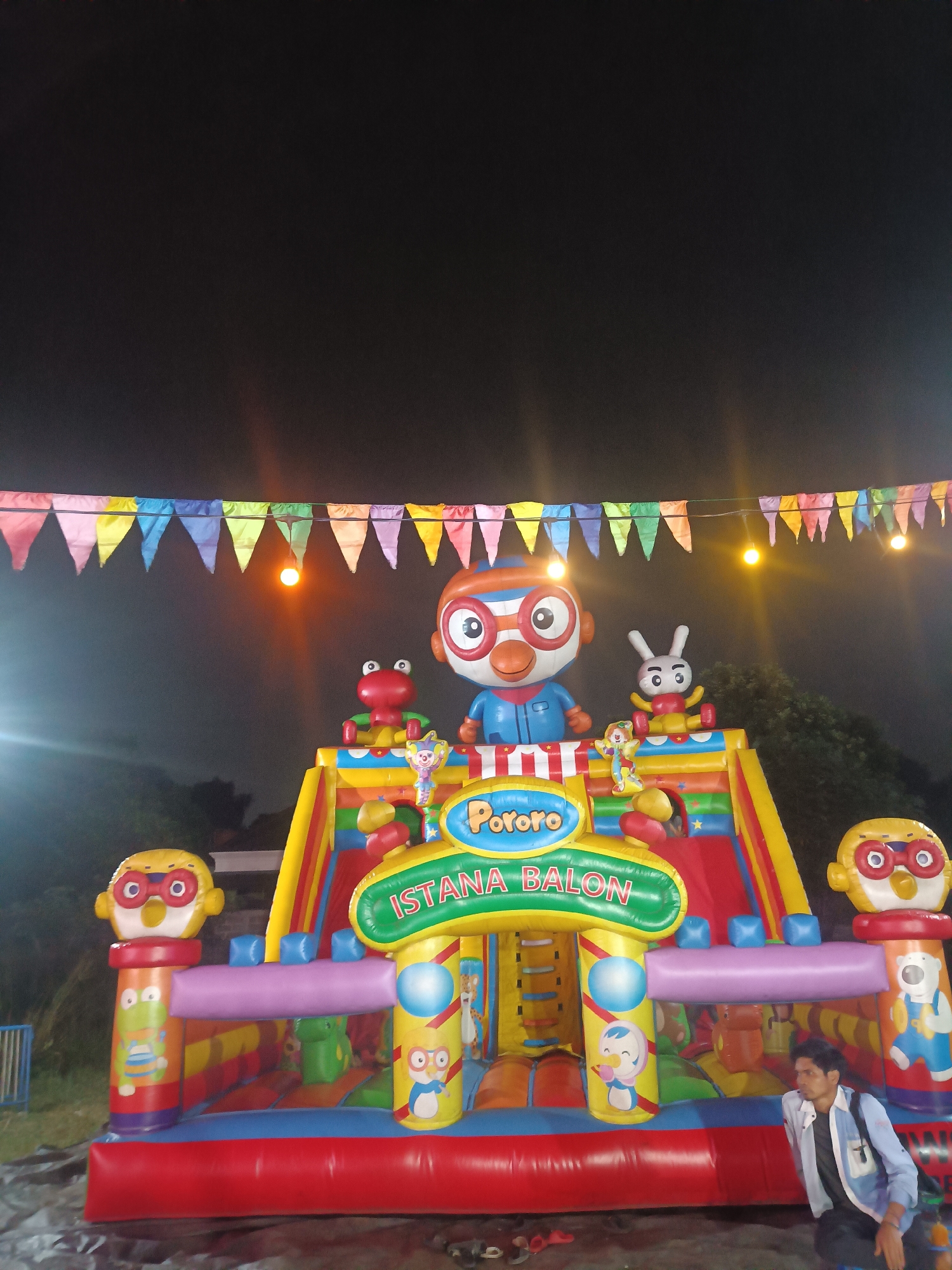 An inflatable playground with characters from the animated series Pororo the Little Penguin is set up at night in a park in Depok, Indonesia. The playground features a large inflatable structure with slides, tunnels, and various cartoon characters. A young man in a light blue jacket is visible in the foreground, observing the playground. Colorful triangular flags are strung across the top of the scene, and lights illuminate the area, casting a warm glow. The background shows trees and a dark sky, suggesting an outdoor event at a local park.