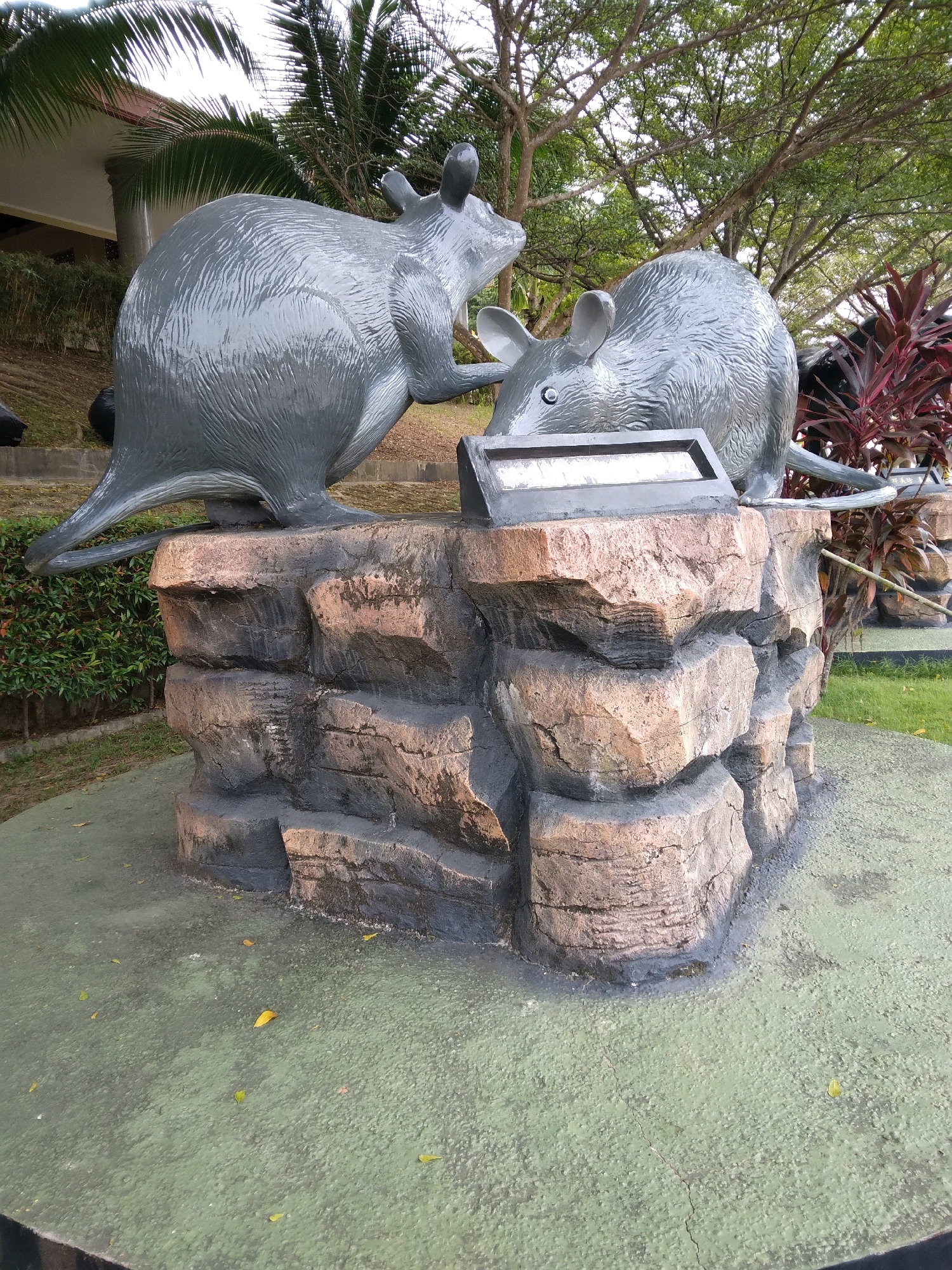 In Batam, Indonesia, two grey, rodent-like sculptures, resembling large rats or possibly kangaroos, are positioned on a textured, rock-like pedestal. The sculpture on the left is larger and faces the right, appearing to be in motion, with its tail extending behind it. The smaller sculpture on the right faces forward, looking downwards. Between the two sculptures, a dark rectangular object with a reflective surface, likely a solar panel or a plaque, is embedded in the pedestal. The pedestal itself is made of stacked, irregular blocks with rough, weathered textures in shades of brown and grey. The sculptures and pedestal are situated on a green, textured surface, possibly a path or ground cover, which is dotted with scattered yellow and green leaves. In the background, lush green foliage and trees create a natural setting, with a hint of a building structure visible on the far left and vibrant red plants on the right. The overall scene suggests an outdoor public space or park.