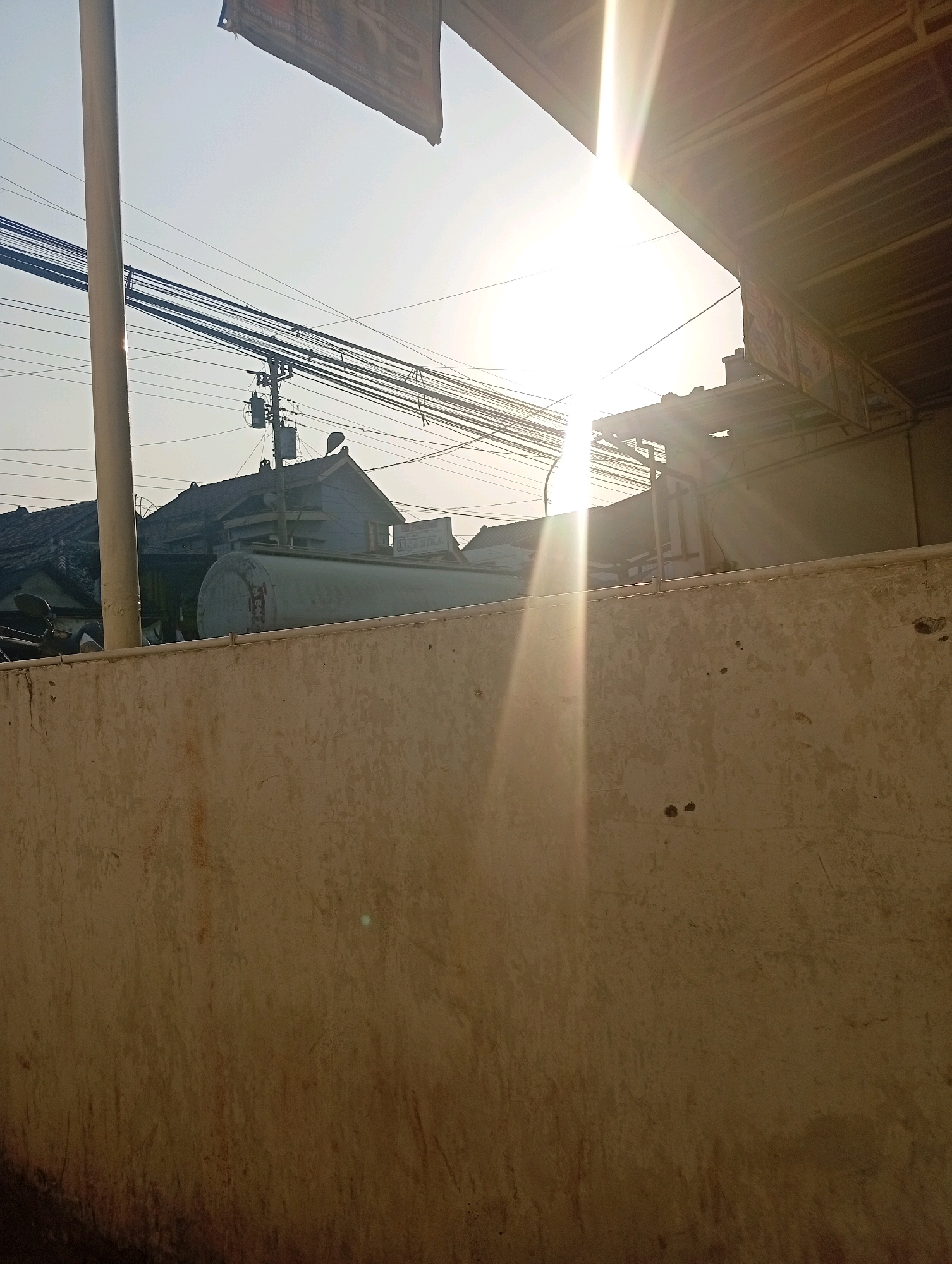 The image depicts a sunlit outdoor scene in Bajingjowo, Indonesia. In the foreground, a textured, light-colored concrete wall stretches across the frame. Behind the wall, a large cylindrical tank is visible, possibly a water or fuel tank. Further in the background, houses with dark roofs are present, along with numerous power lines strung between poles. The sun is bright and high in the sky, creating lens flare and casting strong light across the scene. A portion of an overhang or roof structure is visible in the upper right corner. The overall impression is of an urban or semi-urban environment on a clear day.