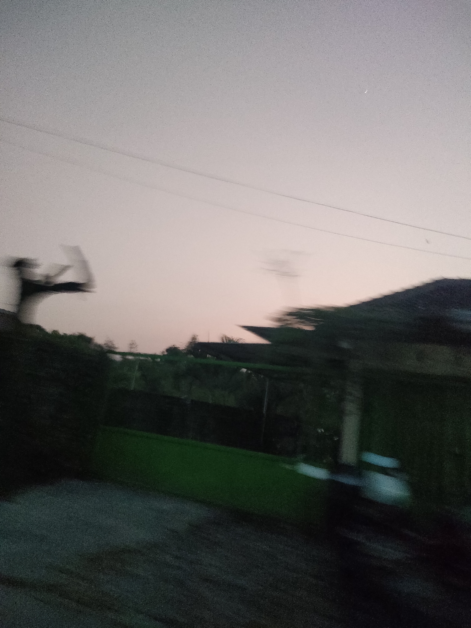 The image is a blurry, motion-filled shot taken at dusk or dawn. The sky is a pale pink and grey gradient. In the foreground, a dark, indistinct object, possibly a person or a vehicle, is blurred due to movement. A green fence runs horizontally across the middle ground, with dark, leafy foliage behind it. Further back, a dark, sloped roof structure is visible. Overhead, two parallel power lines stretch across the frame. A faint crescent moon is visible in the upper right portion of the sky. The scene appears to be in a semi-rural or suburban area, potentially a residential street or a property boundary in Berbah, Indonesia.