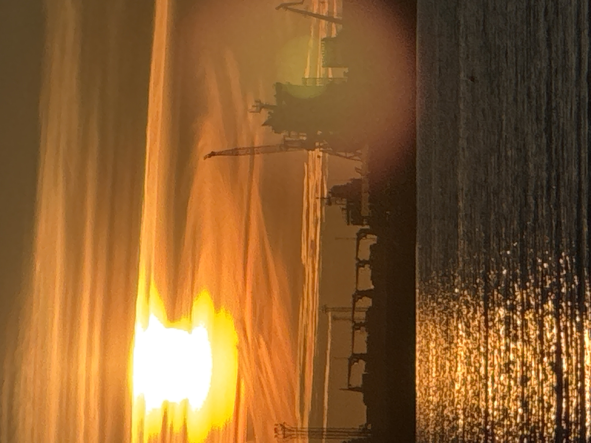 The image depicts a sunset over the port of Tallinn, Estonia. The sun, a bright white disc with a halo, is partially obscured by clouds, casting an orange glow across the sky. Silhouetted against the dramatic sky are industrial cranes and ships, indicative of a busy port. The water in the foreground reflects the warm hues of the sunset, creating a shimmering effect. The overall scene evokes a sense of industrial activity set against the natural beauty of a sunset.