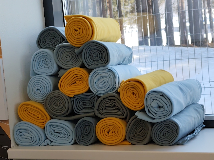 Approximately twenty-seven rolled textiles, consisting of light blue, yellow, and grey fabric, are stacked in an irregular pyramid formation on a white windowsill. The rolls are tightly packed and uniformly shaped. Behind the textiles, a window reveals an outdoor winter scene. This scene includes snow-covered ground and a stand of coniferous trees in the background, partially obscured by a dark-colored metal mesh fence positioned closer to the window. No people or active interactions are present. The location is Chaikovsky, Russia.