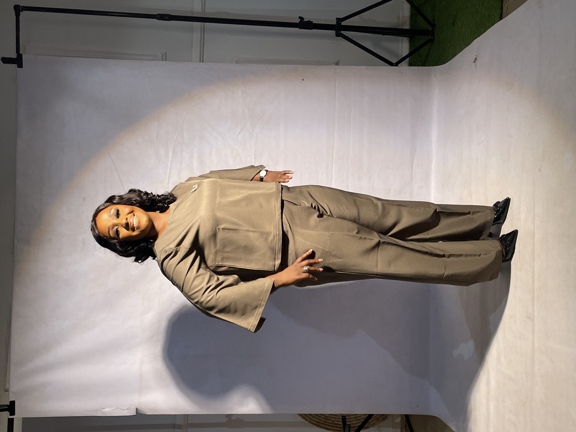 A dark-skinned woman is standing in a room with a white backdrop. She is wearing a tan pantsuit, and black shoes. She is smiling and looking at the camera. There is a black boom stand and a patch of artificial grass visible in the background. The image appears to have been taken in a studio setting. There is no contextual information to verify this location is in the city of Jos, Nigeria.