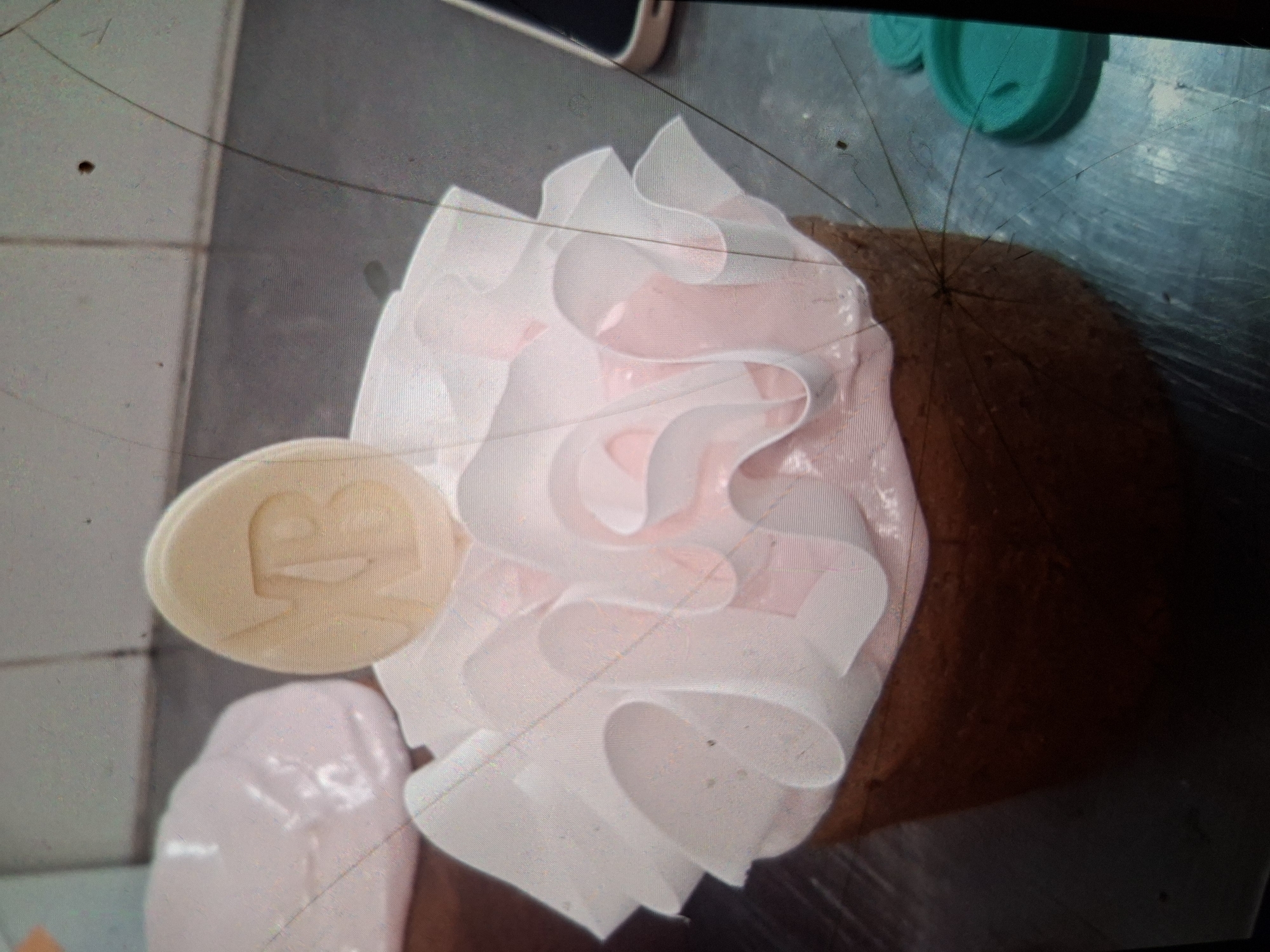 An overhead medium shot captures a cake decorated with pink frosting and white, ruffled paper frills, topped with a light-colored disc embossed with the letters "NB." The cake is positioned on a metallic, reflective surface, possibly a kitchen counter, exhibiting scratches and smudges. To the left of the cake, a partially visible pink blob, possibly meringue or frosting, rests on the surface. In the background, the surface continues, transitioning into a tiled wall. To the right of the cake, two teal lids, likely from containers, are visible. The scene appears to be indoors, possibly in a bakery or kitchen setting in Medvezhyi Ozyora, Russia. No individuals are present in the frame, and the lighting suggests an indoor environment. The image quality is grainy, and several long, dark strands, potentially hairs, are visible across the frame, along with other small dark specks.