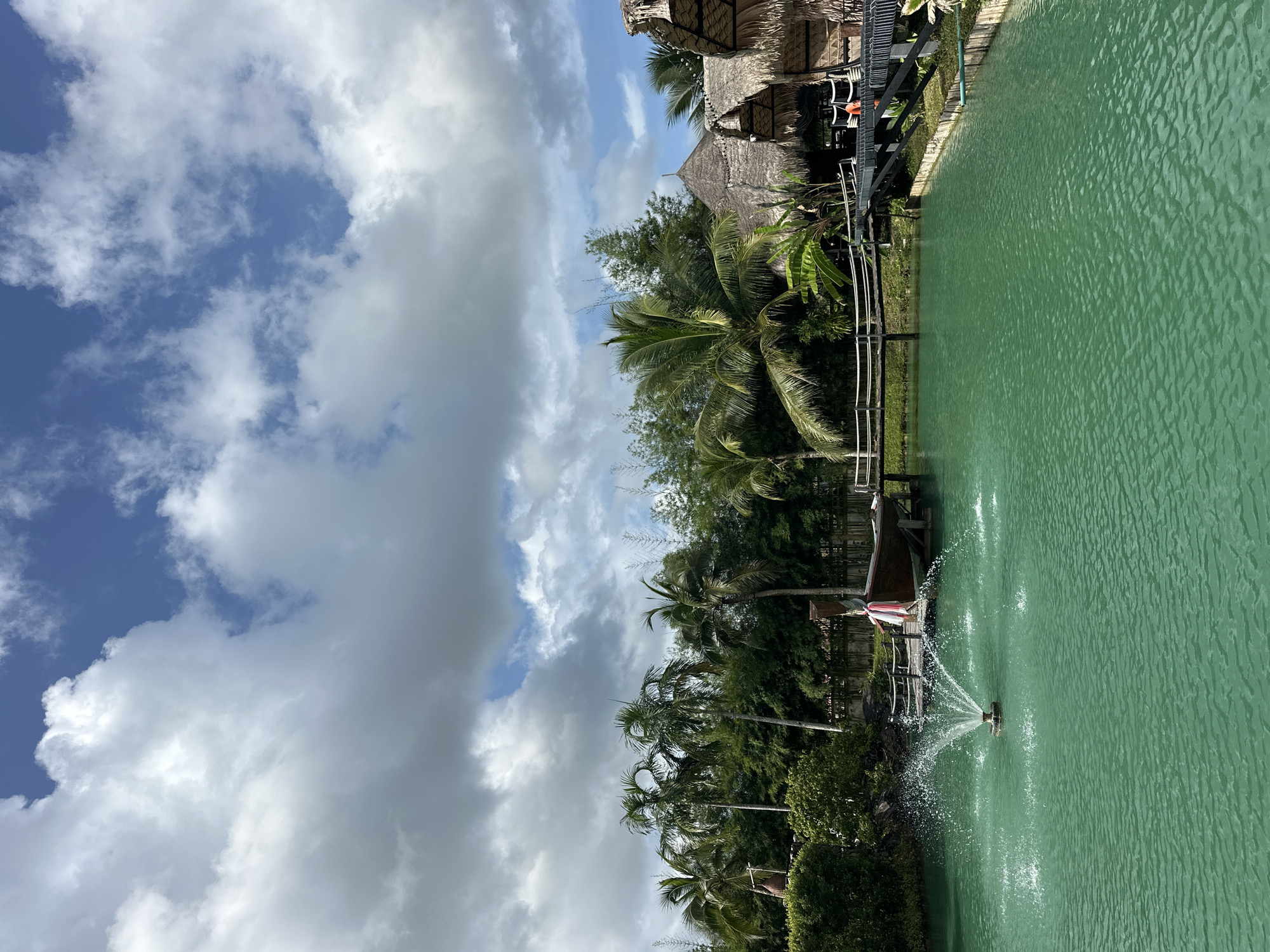 The image depicts a tranquil scene at a location in Tambon Pa Kae Bo Hin, Thailand. A vibrant turquoise body of water occupies the foreground, with a fountain actively spraying water in the left portion. In the mid-ground, a wooden pier extends from the right, leading to a docked wooden boat adorned with a pink and white cloth. Beyond the pier and boat, a lush green landscape is visible, featuring numerous palm trees and dense foliage. To the right, several thatched-roof structures, characteristic of local architecture, are nestled amongst the greenery. The sky above is a mix of blue and white clouds, suggesting a partly cloudy day. The overall ambiance is serene and tropical.
