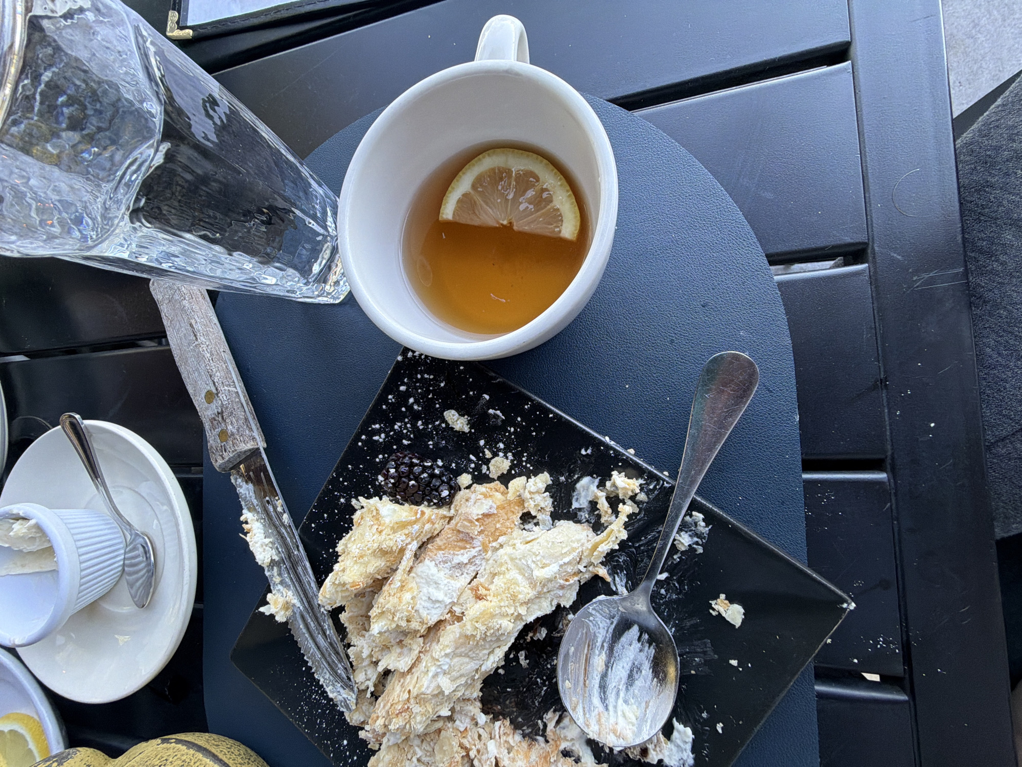 The image shows a table setting, likely outdoors, with various items including a clear glass pitcher, a white mug containing a beverage with a lemon slice, a small dish with a lemon slice, and a black square plate with remnants of a pastry and a blackberry. A knife and spoon, both with traces of cream or icing, are also present. The table is dark, possibly a black plastic or wooden material with a slatted design. The setting appears to be in the city of Glenview, United States of America.