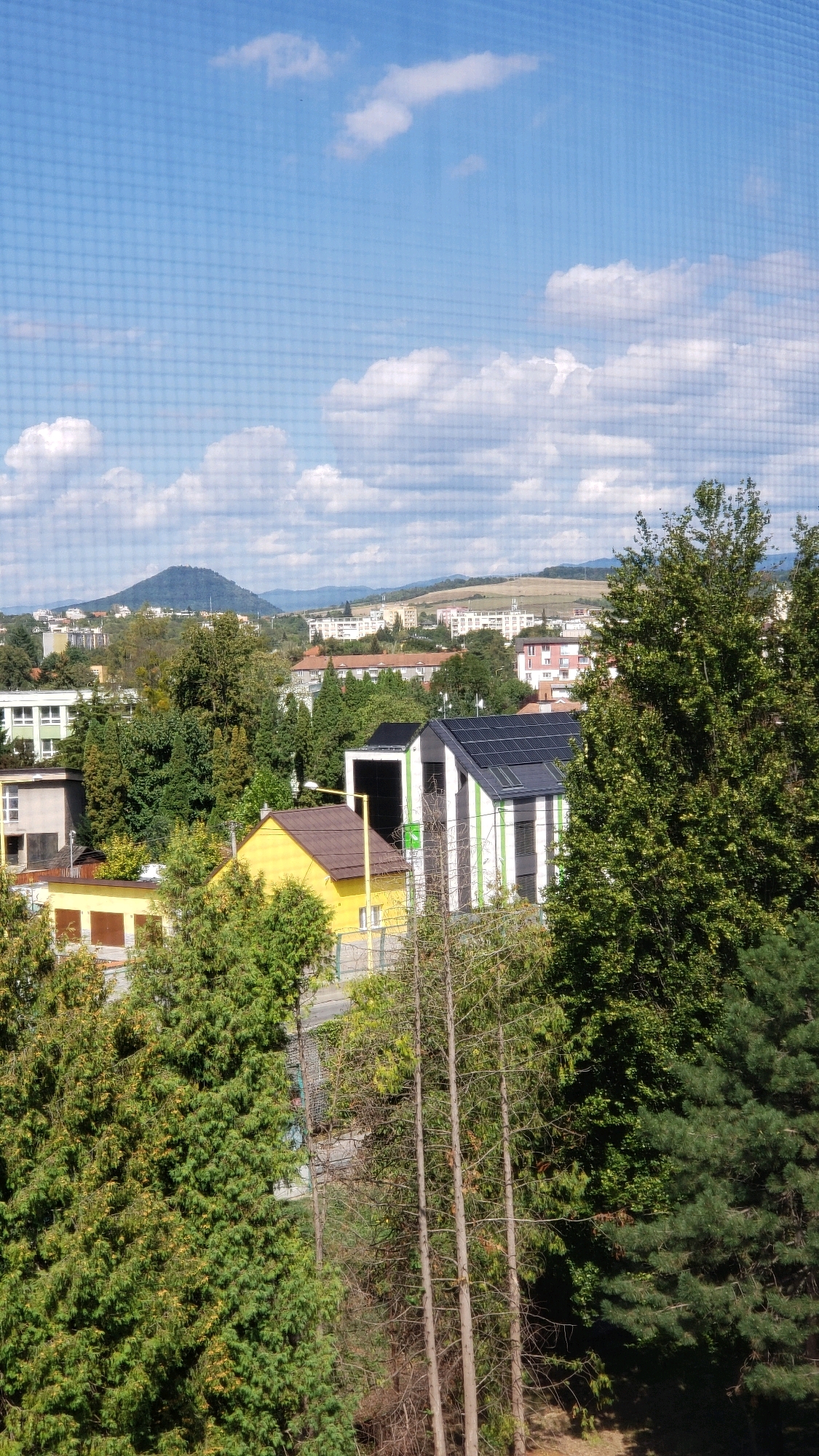 The image depicts a residential area in Prešov, Slovakia, viewed through a grid-like screen, possibly a window screen or netting. In the foreground, lush green trees dominate the scene, with some appearing as evergreens and others as deciduous trees with yellowing leaves. Amongst the trees, several buildings are visible. A vibrant yellow house with a brown roof is situated in the mid-ground. Adjacent to it, a modern building with a white and green striped facade is topped with solar panels. Further in the background, more residential buildings and scattered greenery extend towards a distant, tree-covered mountain. The sky above is blue with scattered white clouds. The overall impression is a snapshot of a sunny day in a suburban or semi-urban setting, characterized by a mix of nature and modern architecture.