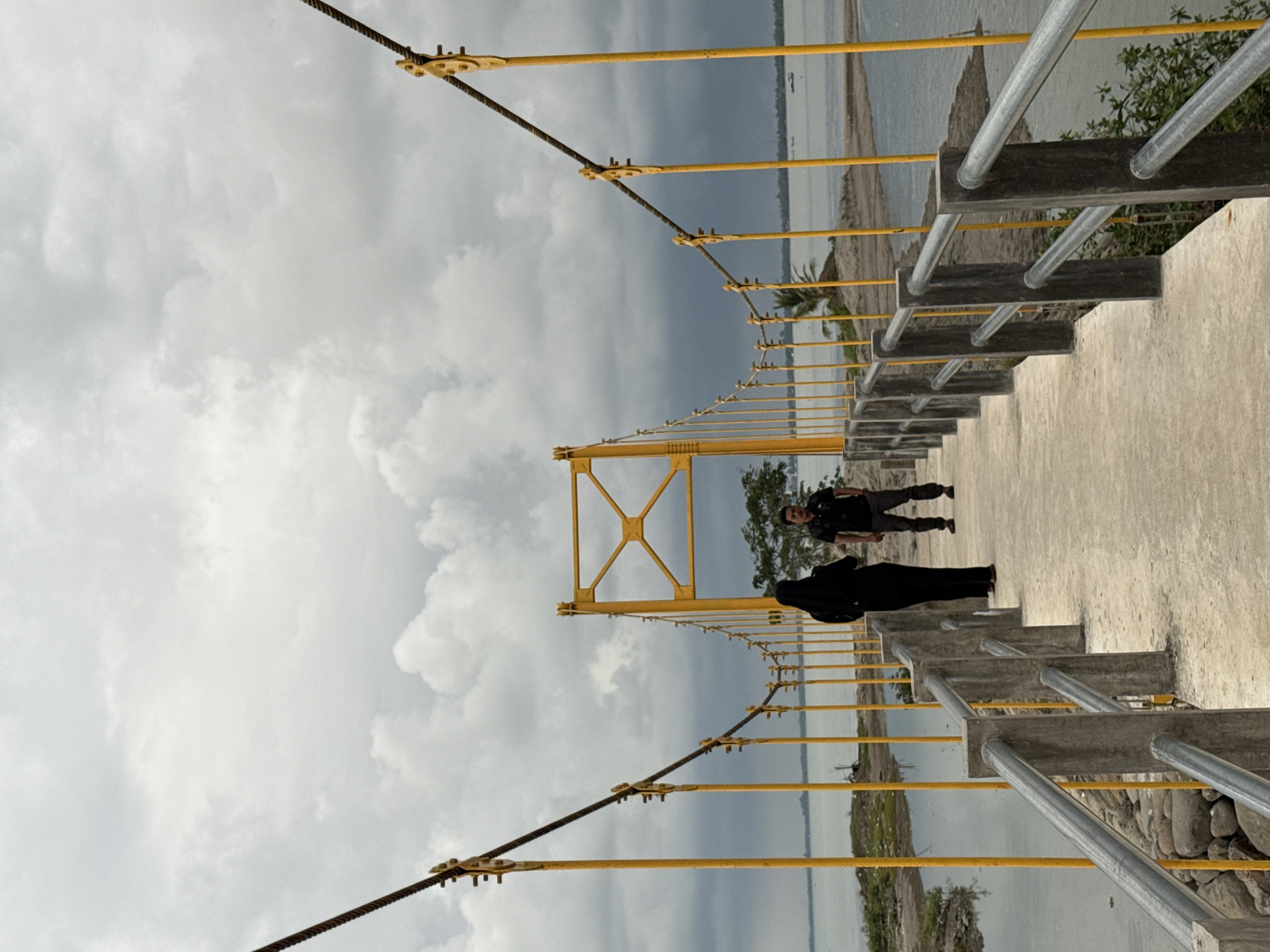 A full, slightly angled shot shows a section of the Krueng Cunda suspension bridge, located in Lhokseumawe, Indonesia, under a cloudy sky.
The bridge's design includes yellow support structures and cables with gray concrete and steel railings. Two individuals are present on the bridge: one dressed in black and another in dark clothing. The environment consists of water and shore visible beyond the bridge's edge.