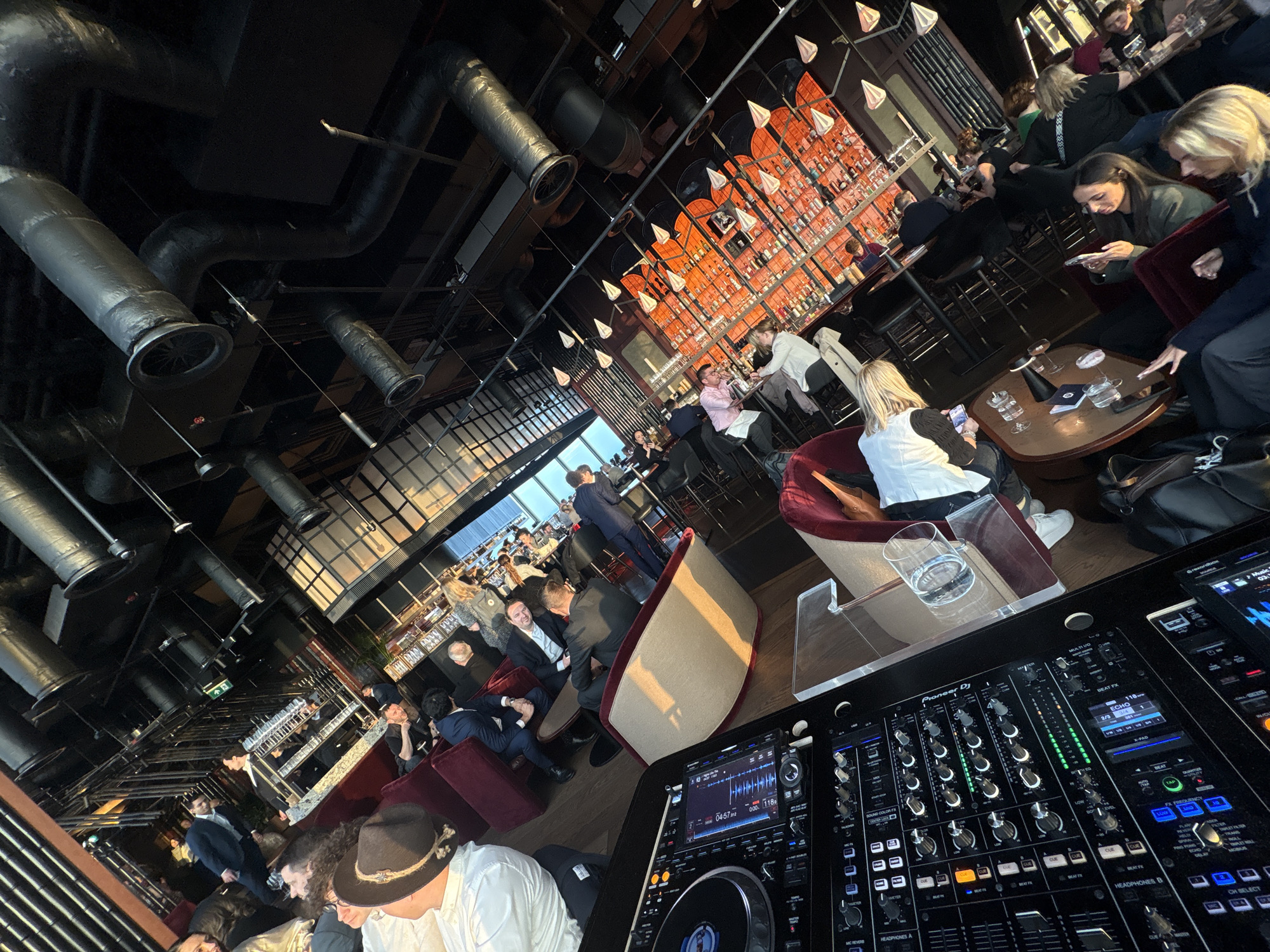 The image shows a crowded bar or lounge setting in London, United Kingdom. The focus is on the foreground, showcasing a DJ setup with a Pioneer DJ mixer and decks. A glass of water sits on a transparent stand next to the equipment. The room is filled with people, some seated in plush chairs and sofas, others standing and mingling. Many are dressed in business attire, suggesting a corporate or after-work event. The background features a well-stocked bar with rows of liquor bottles illuminated by warm lighting. Above, industrial-style ventilation ducts run across the ceiling, adding to the modern atmosphere. The scene appears lively with people interacting, drinking, and socializing.
