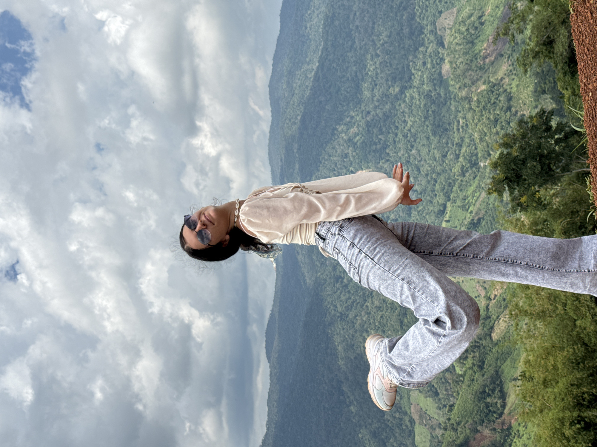 In Nao Sẻ, Vietnam, a young woman stands on a mound of reddish-brown soil, her body angled towards the viewer. She is wearing a light-colored, flowy blouse with long, voluminous sleeves and light blue, high-waisted jeans. Her left leg is bent at the knee and extended backward, her foot shod in a light-colored sneaker. She is looking upwards, her head tilted back, with her eyes closed and a slight smile on her face, wearing round, dark sunglasses. A delicate shell necklace adorns her neck. Her hands are positioned in front of her, palms facing downwards and fingers slightly curled. The background is dominated by a vast expanse of rolling, verdant mountains covered in dense forest, fading into a hazy distance. The sky above is filled with dramatic, gray and white clouds, suggesting an overcast day.