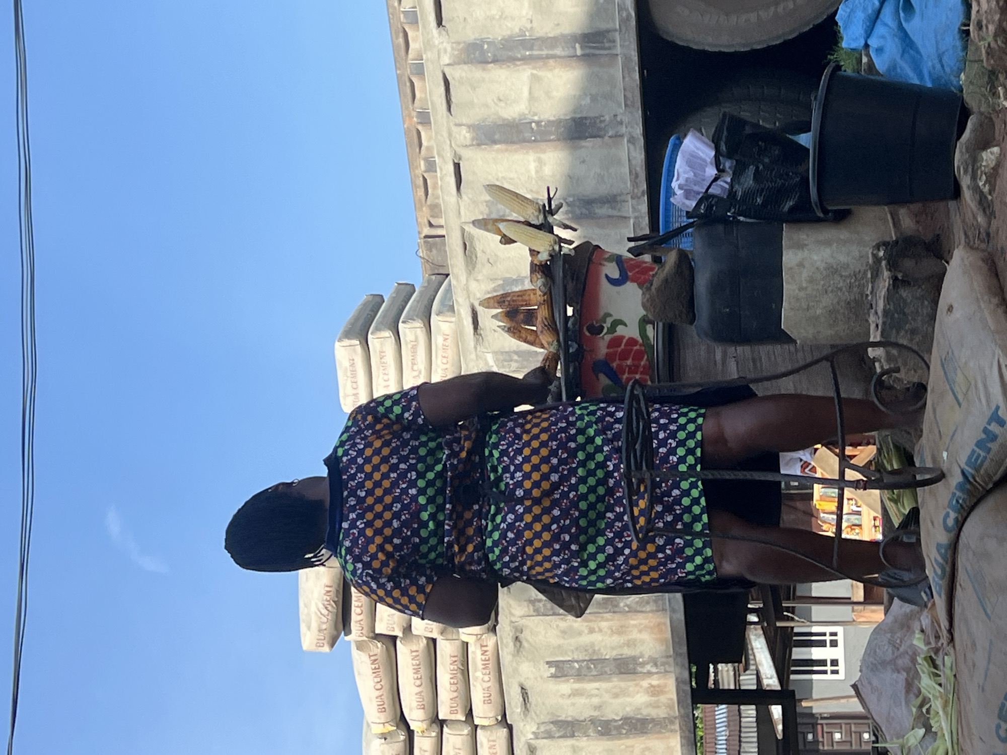 The image shows a person standing beside stacks of cement bags. The person is wearing a colorful, patterned dress and has dark hair. They appear to be in an outdoor setting, possibly a market or construction site, given the presence of the cement bags and the blue sky above.
In the background, there are more stacks of what appear to be bags of cement, with the text "BUA CEMENT" visible on them. There are also some miscellaneous objects, including buckets and what looks like dried corn. The lighting suggests it is daytime, and the clear blue sky indicates fair weather. The overall scene conveys a sense of commerce or perhaps preparation for construction work. The person's posture doesn't strongly suggest a particular emotion, appearing to be in a neutral stance. The location is identified as Nkpor, Nigeria.