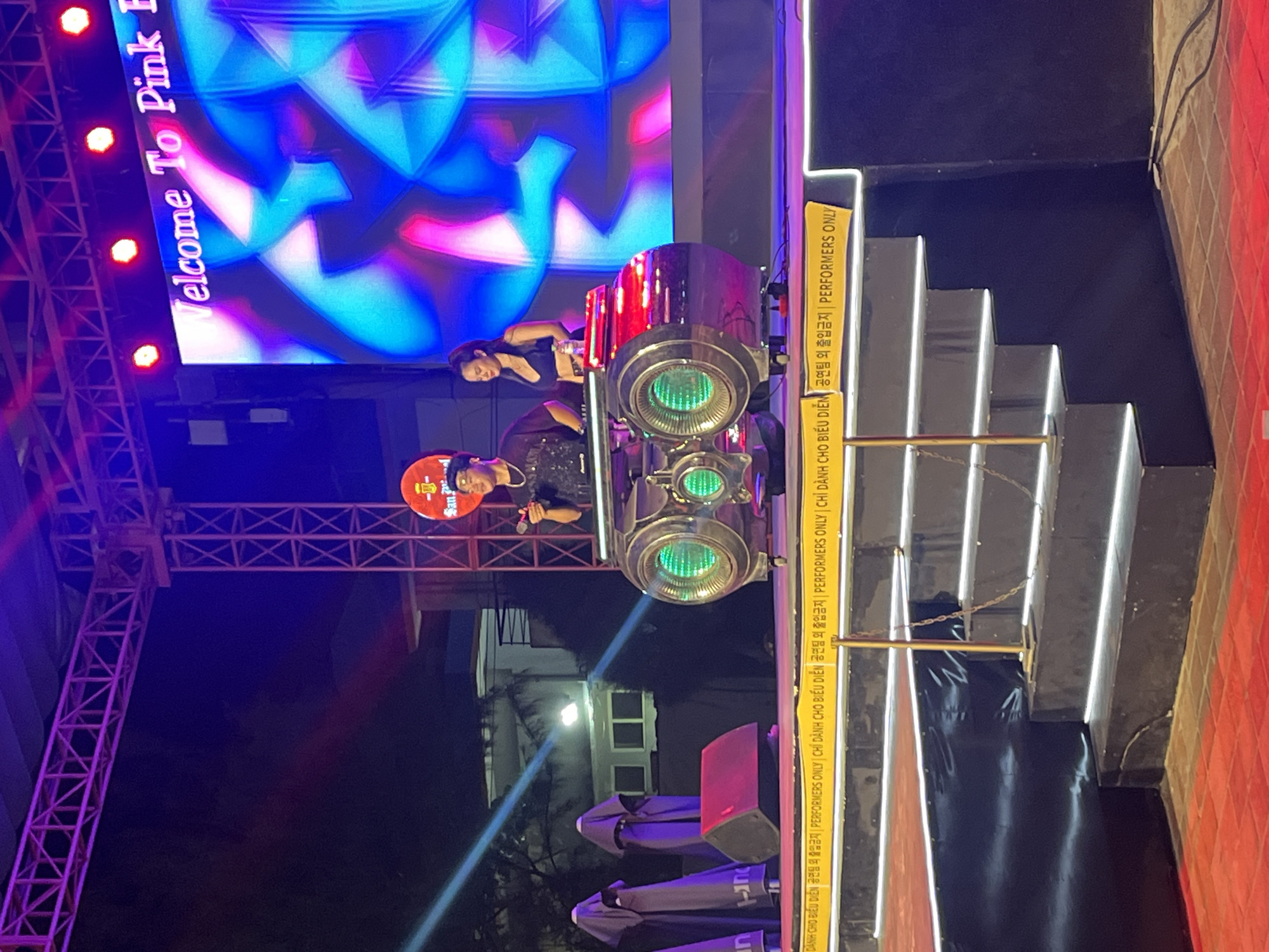 The image depicts an outdoor nighttime event, likely a performance or concert, in Đà Nẵng, Vietnam. The scene is dominated by vibrant purple and blue stage lighting illuminating a large LED screen displaying abstract geometric patterns.
In the foreground, two individuals are partially visible. One, wearing a red cap, is holding what appears to be a microphone and looking towards the left. Above and behind them, another person in a black top is leaning forward, possibly interacting with equipment or a performer. A large, cylindrical object with green glowing lights is prominently featured, suggesting it might be a stage prop, lighting equipment, or part of a performance setup.
A yellow banner runs vertically along the right side of the frame, featuring text in Vietnamese and English. The Vietnamese text, "DÀNH CHO ĐIỀU DIỄN" and "Công ty biểu diễn," translates to "For performers" and "Performance company," respectively. The English text clearly states "PERFORMERS ONLY." This indicates a restricted area for performers.
Behind the stage area, a set of illuminated stairs leads upwards, with a metal railing. The overall ambiance is energetic and festive, characteristic of a live entertainment venue at night. The presence of outdoor seating with folded umbrellas in the background suggests a public gathering space.