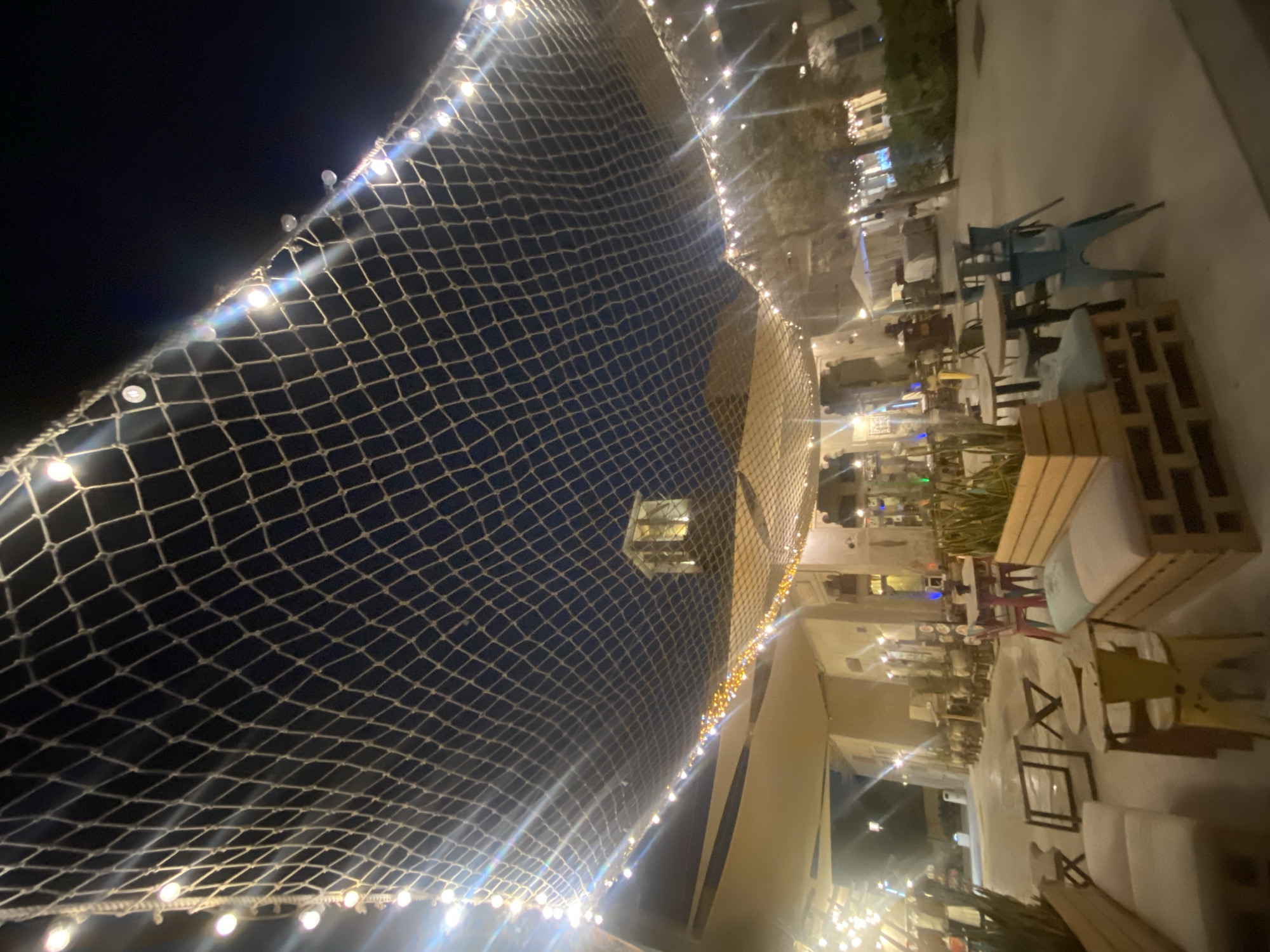 The image depicts an outdoor seating area at night, possibly a restaurant or cafe, in Deira, United Arab Emirates. A large net structure, strung with numerous small lights, is stretched across the upper portion of the frame, creating a canopy effect against the dark night sky. Below the net, more strings of lights are visible, illuminating the scene.
The area features various seating arrangements, including wooden benches with cushions, metal chairs in different colors (some blue, some pink, some yellow), and small tables. Some tables are round, while others are square or have an abstract design. Potted plants are strategically placed, adding a touch of greenery. In the background, there are buildings with traditional architectural elements, and a tower-like structure with a lit top is visible. The overall atmosphere is illuminated by artificial lighting, giving the scene a warm and inviting ambiance despite the darkness of the night. No people are clearly visible in the image, but the setup suggests a place intended for patrons.
