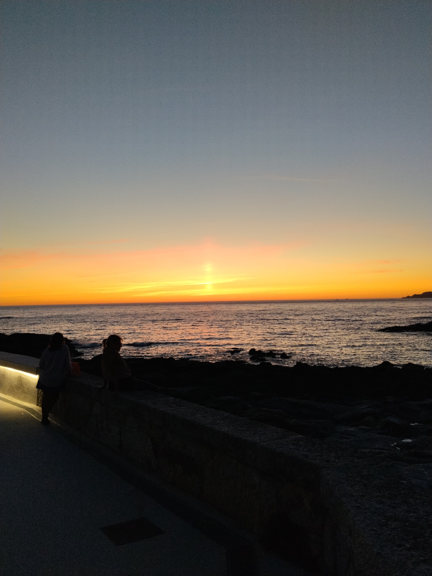 A serene sunset is captured over the Atlantic Ocean in Baiona, Spain. Two figures are silhouetted against the warm hues of the sky, which transitions from a deep blue at the top to vibrant oranges and yellows near the horizon. They are seated on a stone wall overlooking the water. The foreground features a paved walkway illuminated by a soft, upward-facing light. Dark, rocky terrain lines the edge of the water, with the gentle waves reflecting the colorful sky.