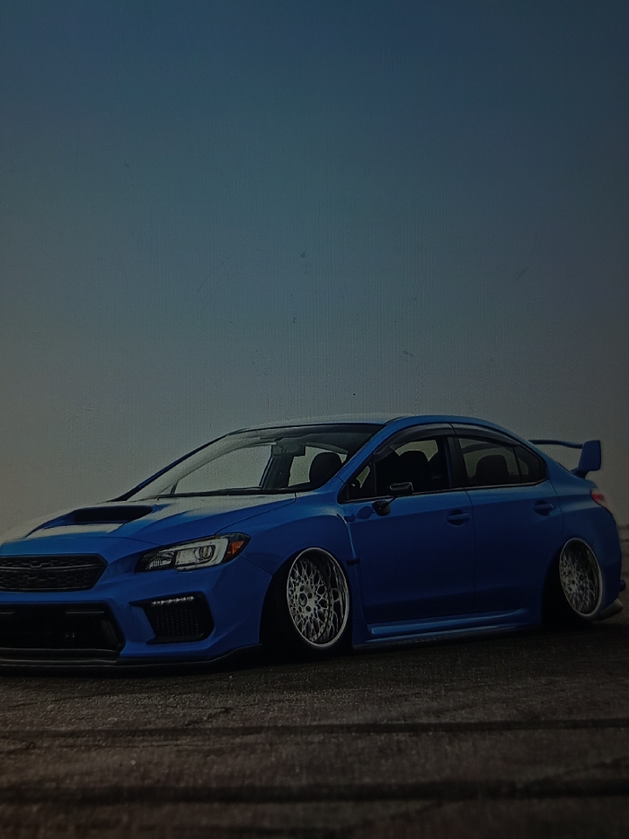 A modified dark blue Subaru WRX sedan is stationary, positioned at a low ride height on a textured, dark grey paved surface. The vehicle is viewed from a three-quarter front perspective. It features extreme negative camber on its polished, multi-spoke aftermarket wheels. Visible modifications include a prominent hood scoop, a custom front bumper with integrated grilles, side skirts, and a rear spoiler. The background is a uniform light blue-grey gradient sky, devoid of any discernible horizon line or structural elements. No individuals or active interactions are present in the scene. The location is Reutlingen, Germany.