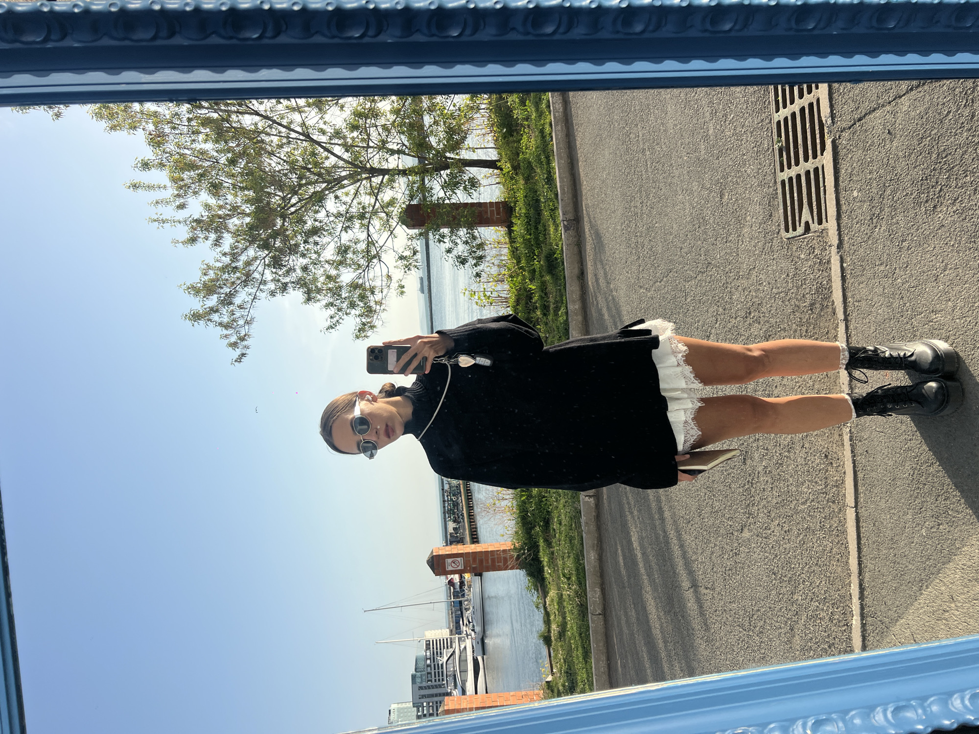 A photograph shows a woman in a black dress with a white lace hem and black boots taking a selfie in a blue-framed mirror. The mirror reflects a cityscape with a harbor containing several yachts, brick pillars along a waterfront, a tree, and a road with a storm drain. The woman is wearing sunglasses and holding a phone. The scene appears to be outdoors on a sunny day. There is no indication of the location being Vladivostok, Russia.