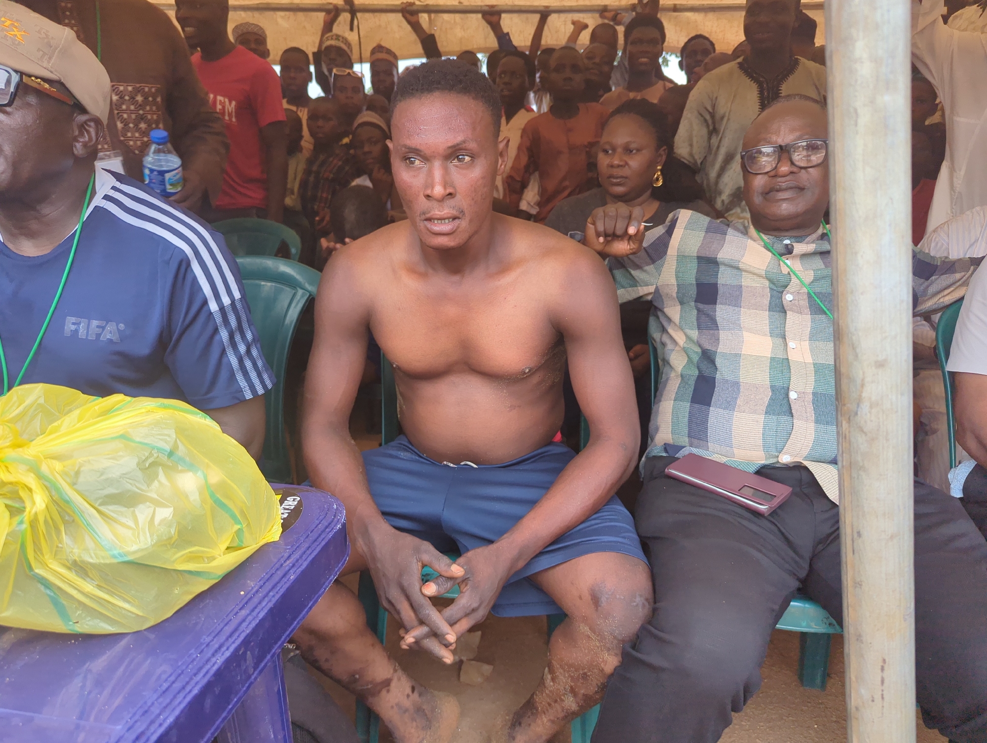 A man is seated shirtless and wearing shorts with dirt on his legs. He is flanked by two men: one in a FIFA-branded shirt and the other in a plaid shirt. A crowd of people is visible in the background. The setting appears to be outdoors under a tent, with a blue plastic table and a yellow plastic bag to the left. The location is Kafanchan A, Nigeria.