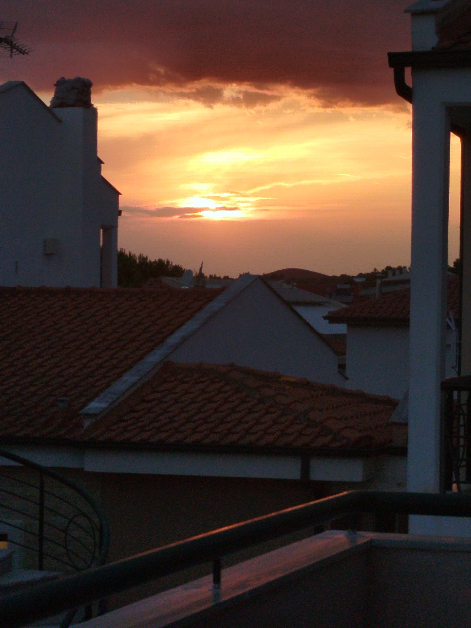 The image depicts a sunset over a city skyline. The sky is a dramatic blend of deep purples, oranges, and yellows, with clouds casting shadows. In the foreground, the tiled roofs of several buildings are visible, along with architectural elements such as chimneys and portions of walls. A metal railing and a spiraling structure suggest a balcony or terrace. The scene appears to be an outdoor urban environment at dusk.