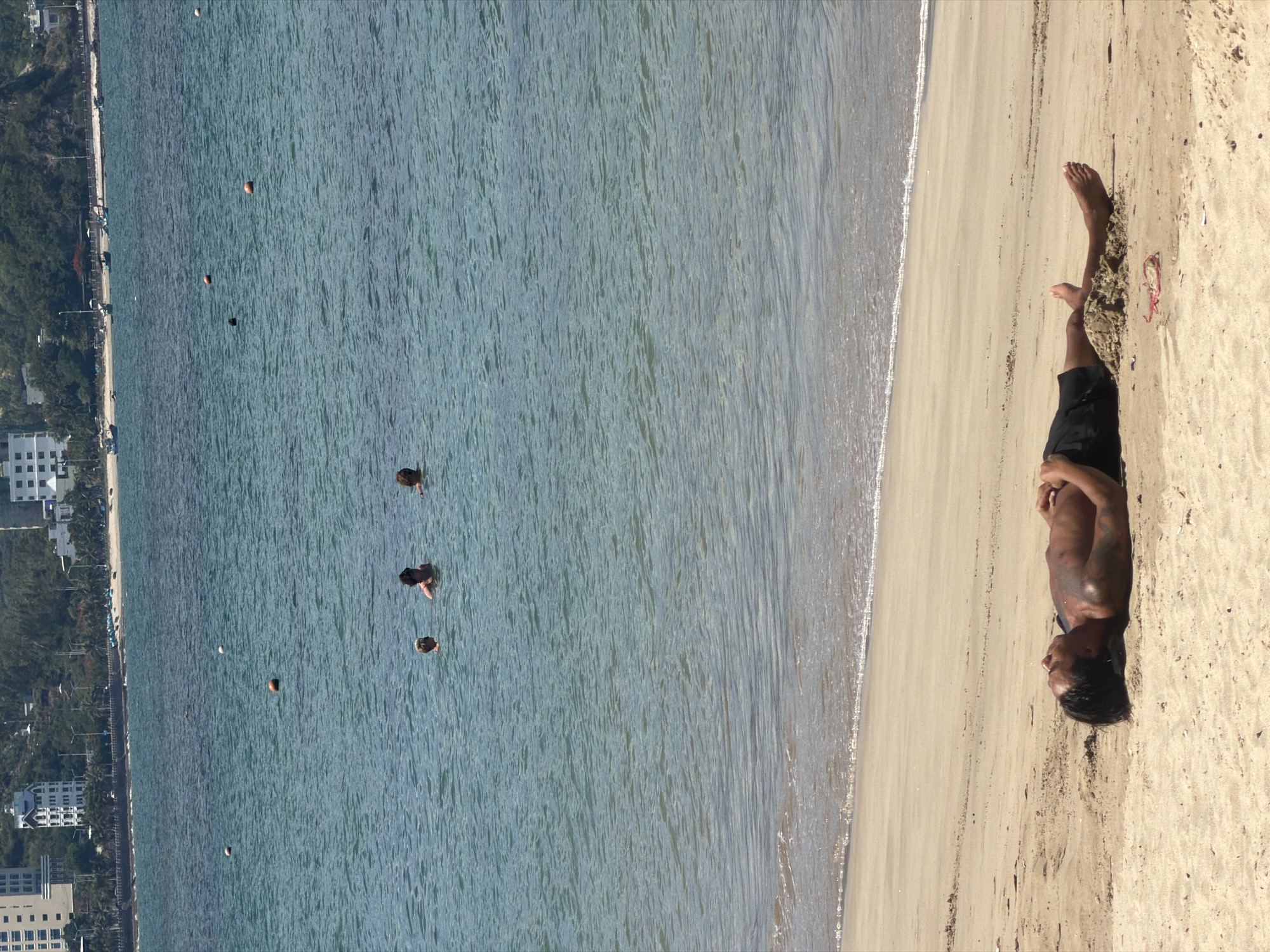 A man lies on his back on a sandy beach, with his head resting on the sand. He is wearing black swim trunks. The sea is calm and blue, with several buoys visible in the distance. A few people are swimming in the water. In the background, buildings and trees can be seen along the coastline of Nha Trang, Vietnam.