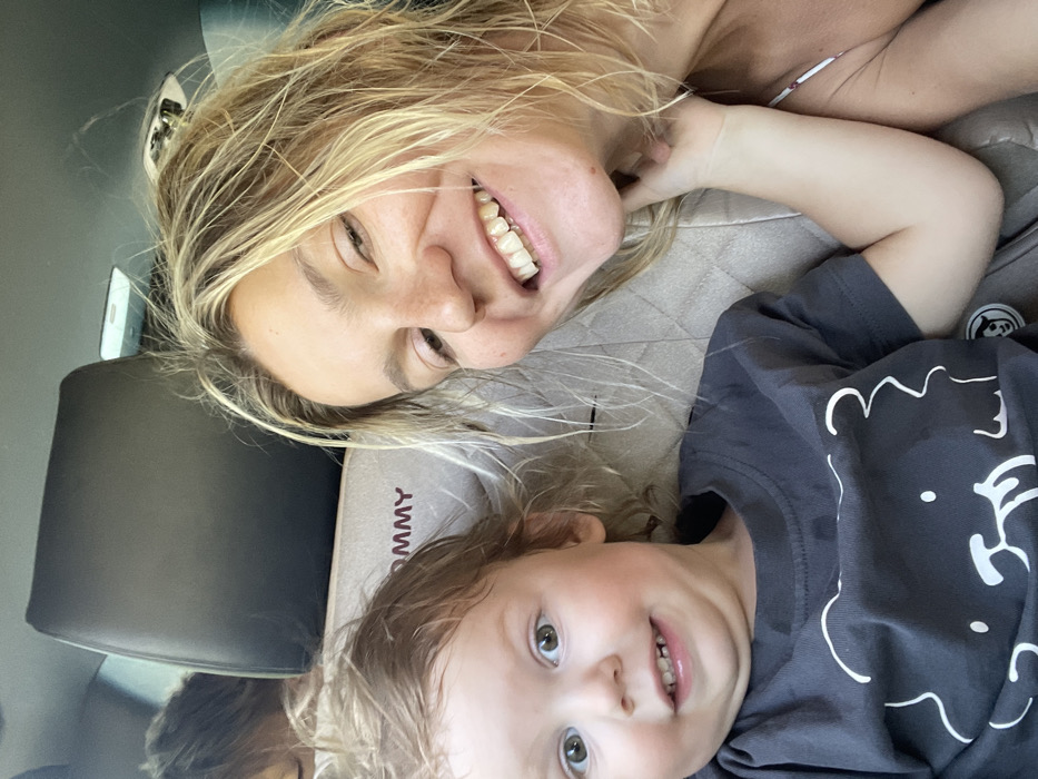 The image depicts two primary subjects inside a vehicle, identified as being in Sukko, Russia. In the foreground on the right, an adult female with unkempt blond hair and a smiling expression is visible, appearing to take a selfie. Her right hand is positioned near her neck. To her left and slightly lower, a young child with light brown, curly hair and wide eyes is seated in a beige quilted car seat, also smiling and looking towards the camera. The car seat's headrest features "TOMMY" embroidered in red capital letters. The child wears a dark grey t-shirt adorned with a white outlined bear design. The child's left hand is visible, resting on or near the adult female's neck. In the background, partially obscured by a dark vehicle headrest, the crown of a third individual's head with dark hair is visible. The vehicle's interior features include a light-colored ceiling with a light fixture. The scene is illuminated by natural light, indicating daytime.