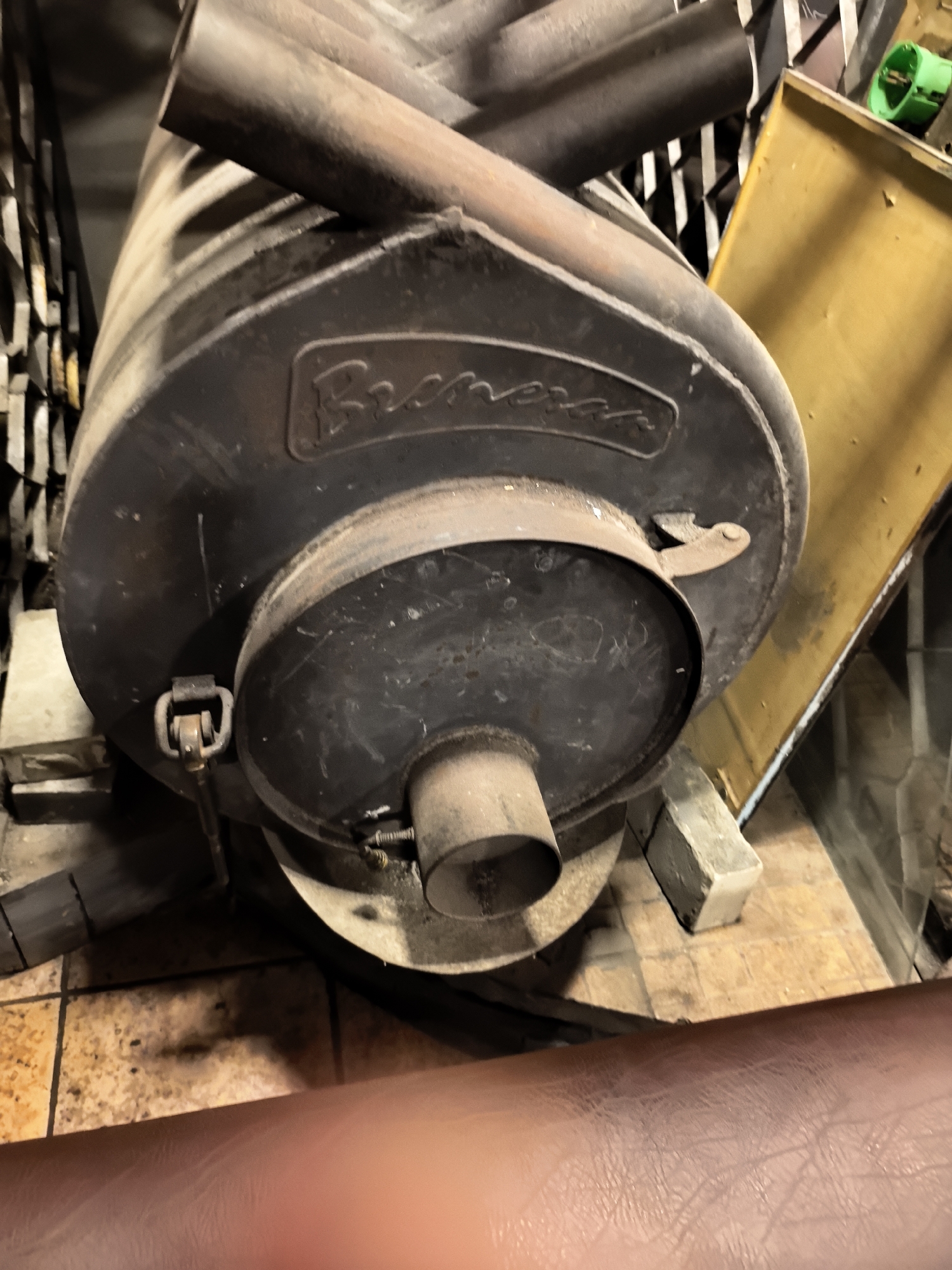 A close-up shot reveals a round, black metal wood-burning stove, likely a "Brezar" model, with associated exhaust pipes on top. The stove appears to be in a utilitarian setting, resting on what looks like concrete blocks on a tile floor. A yellow panel leans against the stove. The lighting is dim, highlighting the worn texture of the stove and suggesting an indoor environment. The bottom portion of a brown leather object is present in the foreground.
There is no data to support this image being taken in Kamenka, Russia.