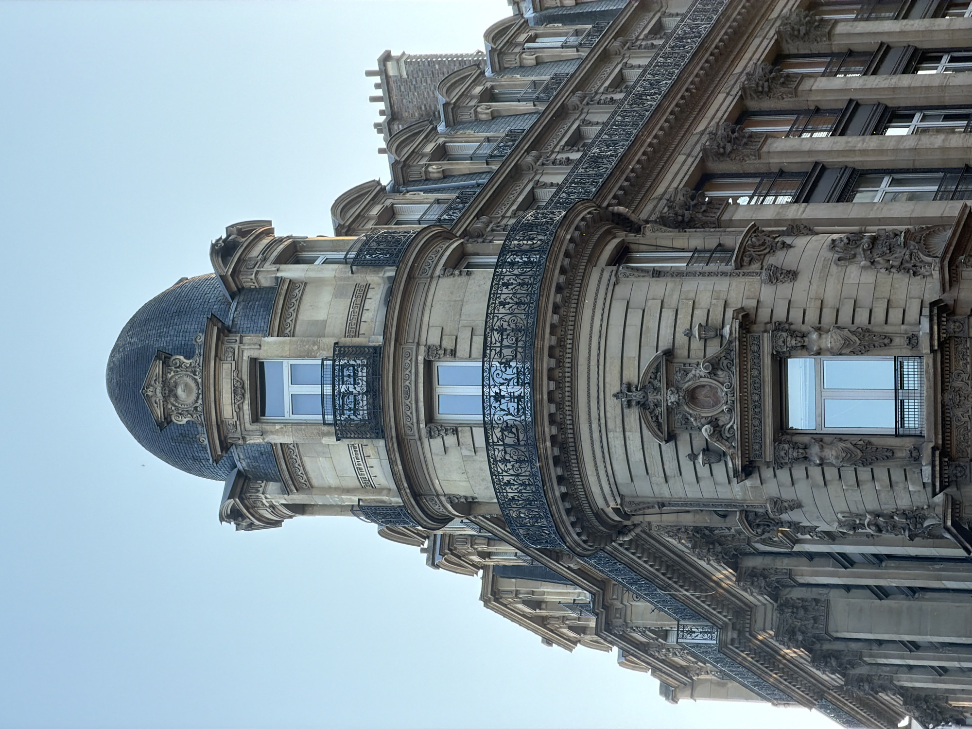 The image captures an upward-angled view of an ornate, multi-story building facade in Paris, France, under a clear, bright blue sky. The architecture is characterized by its grand scale and intricate detailing, typical of Haussmannian or Belle Époque styles.
The building is constructed from light-colored stone, adorned with numerous decorative carvings, including classical motifs, relief panels, and sculptural elements. A prominent feature is a large, dark, domed roof, likely made of slate or zinc, which caps a rounded corner of the building. This curved section also incorporates several windows and gracefully arcing wrought iron balconies with elaborate, scrolled patterns. Additional wrought iron railings and balconies extend along the straighter sections of the facade.
Multiple windows with white frames are visible across the different stories. The strong daylight casts distinct shadows and highlights, emphasizing the depth and three-dimensional nature of the architectural details. No people are visible, and there is no discernible activity taking place. The scene conveys the historical elegance and grandeur of Parisian urban architecture. No visible text is present in the image.