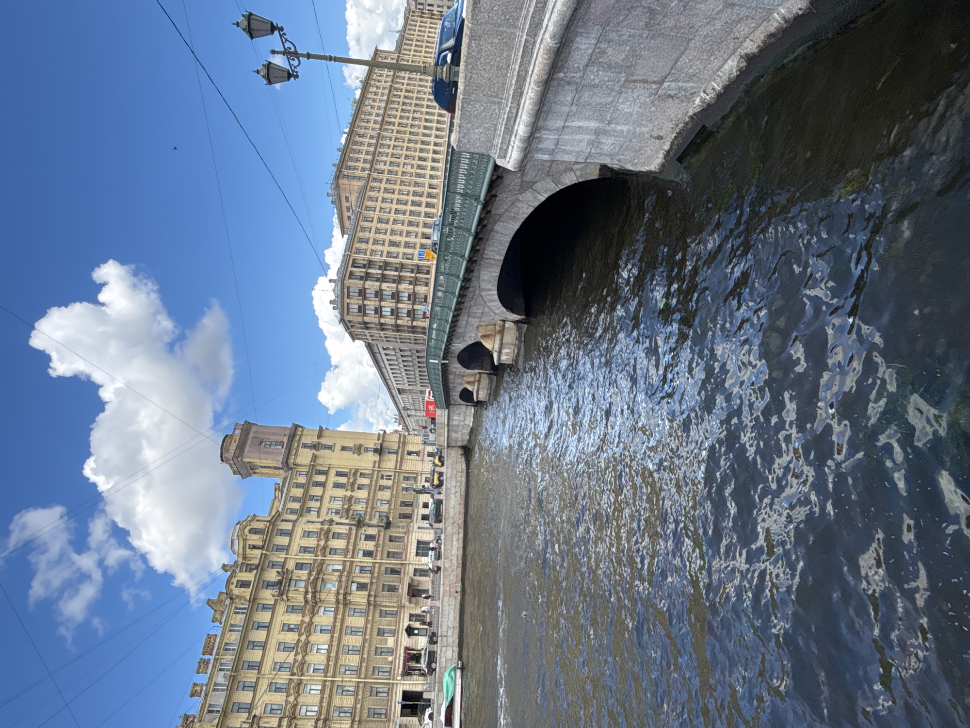 A sunny day in Saint Petersburg, Russia, features a view of a stone bridge spanning a dark, rippling river. The bridge, with its green railing, has two archways visible. On the left bank, a light yellow apartment building with many windows and architectural details is prominent. Several cars and a boat are parked along the embankment. Across the river, another large, light-colored apartment building stretches along the bank. A classic street lamp with two lights stands on the right side of the image. The sky is a bright blue with scattered white clouds, and the water reflects the sky and surrounding buildings.