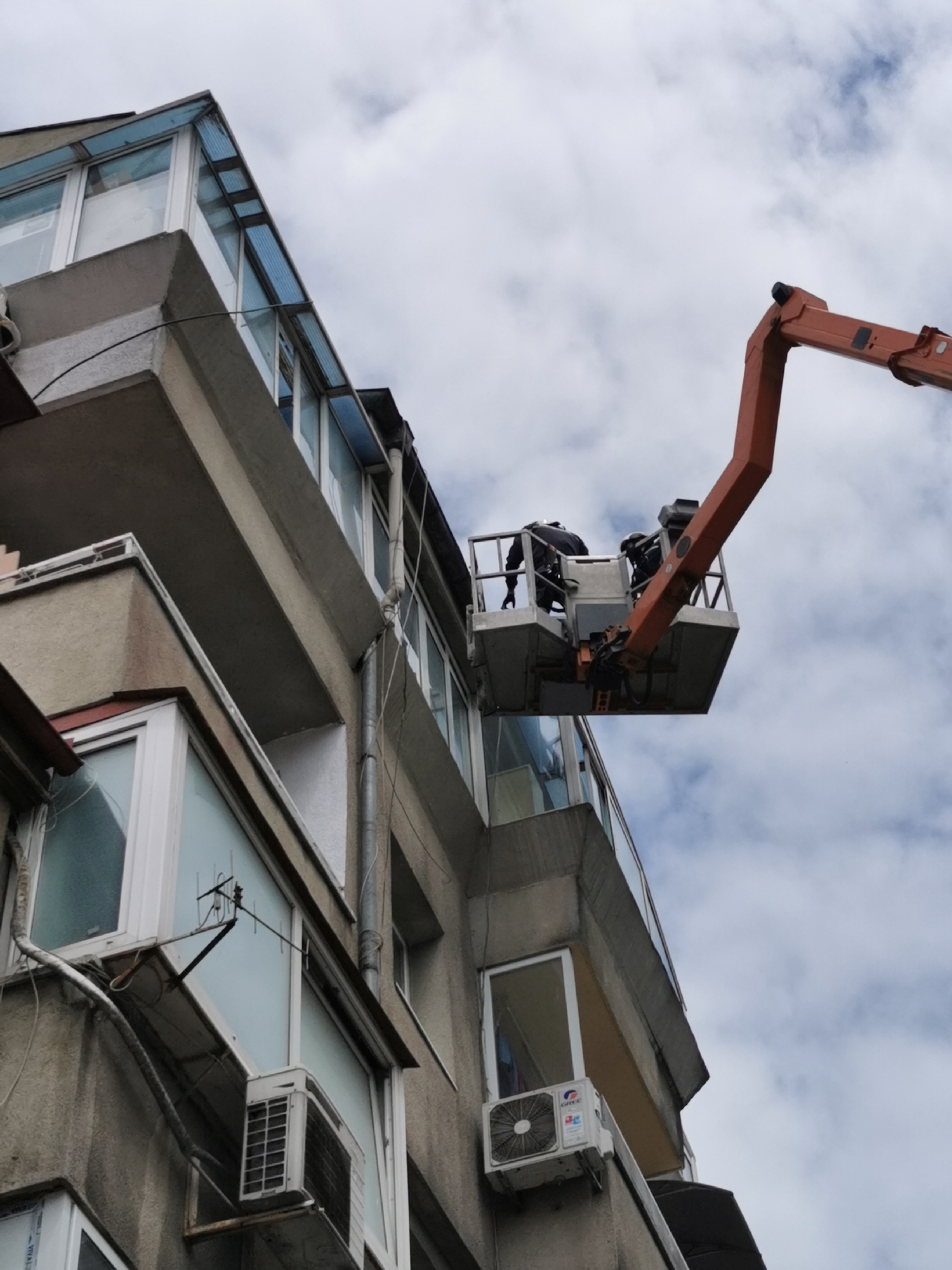 In Varna, Bulgaria, an elevated work platform with two workers in safety gear is positioned near a building's upper floors. The building has several balconies with glass enclosures, windows, air conditioning units, and a drainpipe. The sky is overcast with clouds.