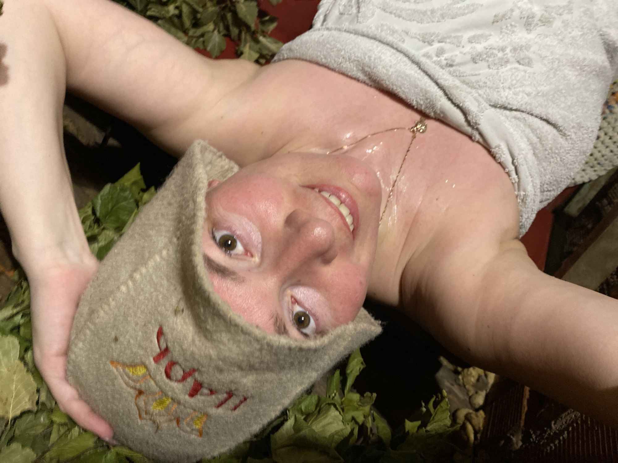 A fair-skinned person wearing a sauna hat with Cyrillic script and leaf designs is lying on their back amidst scattered green leaves. The person is partly covered by a light gray towel, and a gold chain necklace is visible on their neck. The person is smiling and looking towards the camera. The scene appears to be indoors, with a dark background. Based on the hat inscription and the prompt information, this location is likely Noviy, Russia.