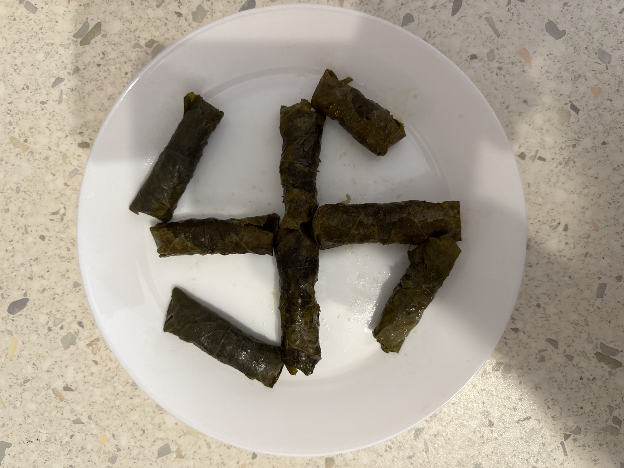 The image shows a white plate containing dolmades arranged in the shape of a swastika. The plate is on a speckled countertop. There are no people, actions, or interactions depicted. There is no information in the image that would allow for the location to be identified as Milan, Italy.