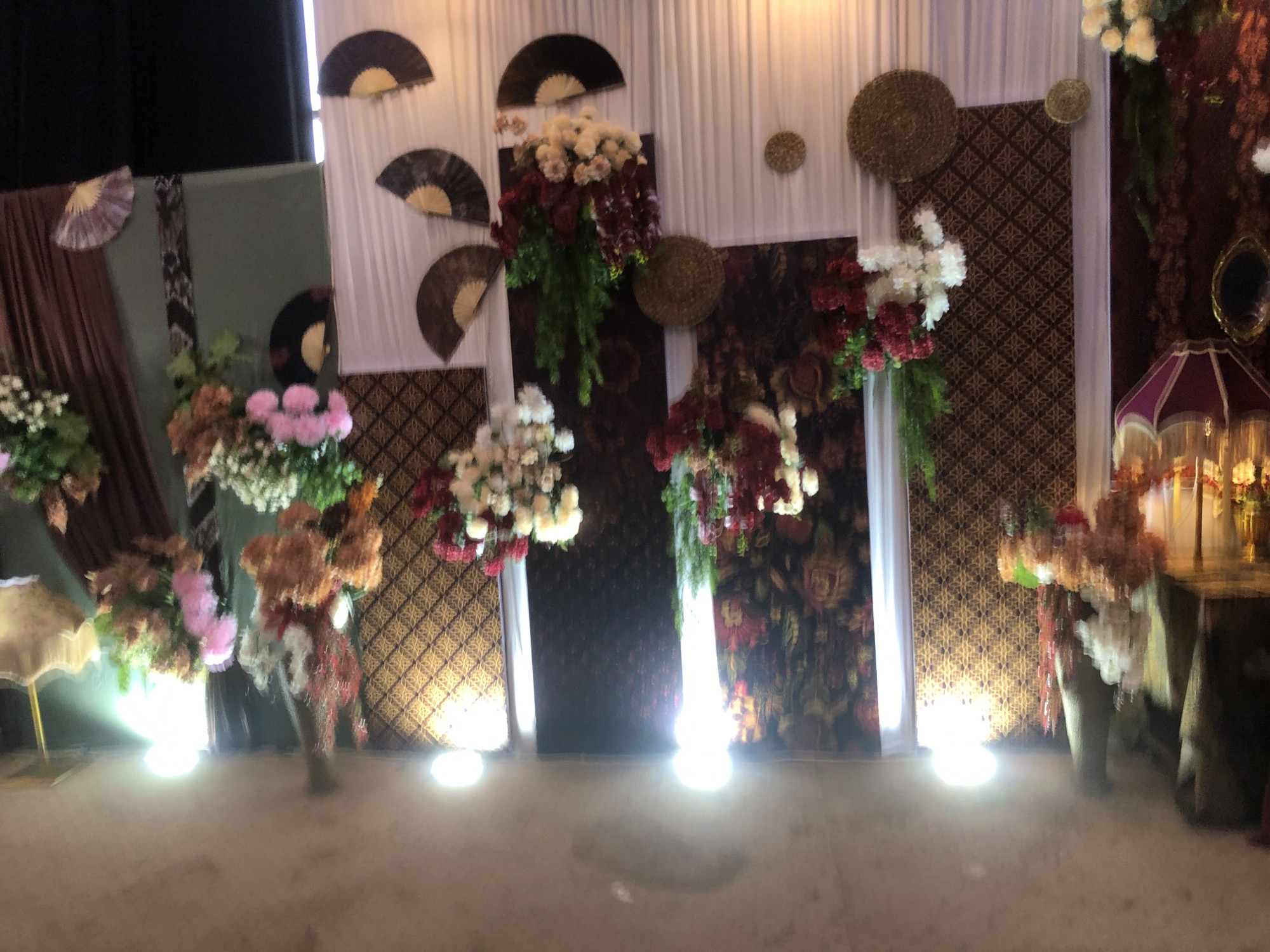 The image captures an indoor setting adorned with floral arrangements and decorative fans, likely for an event. The backdrop features draped white curtains and patterned fabric panels. Several bouquets of flowers, predominantly in shades of red, white, and pink, are prominently displayed. Decorative fans, some of which appear to be made of natural materials, are affixed to the wall. The lighting suggests an evening event. There are no people visible in the image. This scene is consistent with a wedding or celebration venue, which can be found throughout Lhokseumawe, Indonesia.