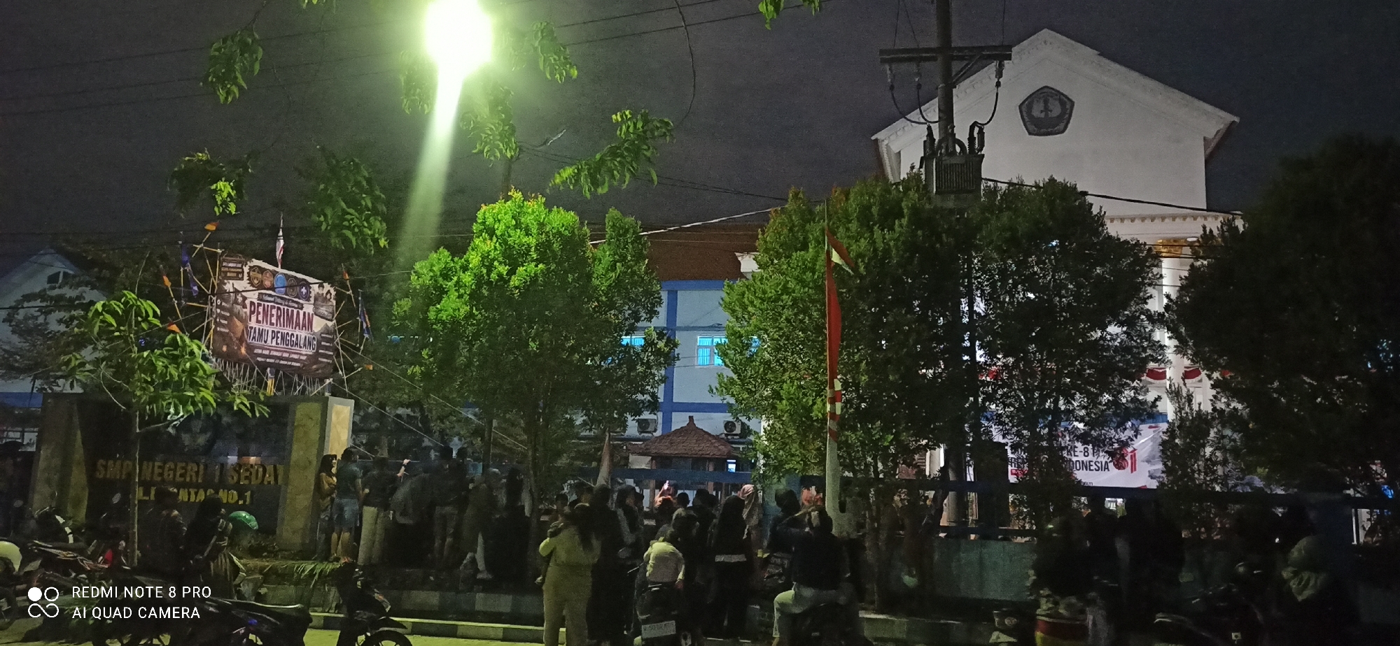 At night in Sedati Agung, Indonesia, a group of people are gathered in front of SMP Negeri 1 Sedati. A large banner with Indonesian text, including "PENERIMAAN TAMU PENGGALANG," is prominently displayed. Trees are scattered throughout the scene, some illuminated by a bright overhead light. In the background, a building with blue-tinted windows and a white facade is visible. Several motorcycles are parked in the foreground. The overall atmosphere appears to be one of a public gathering or event.
