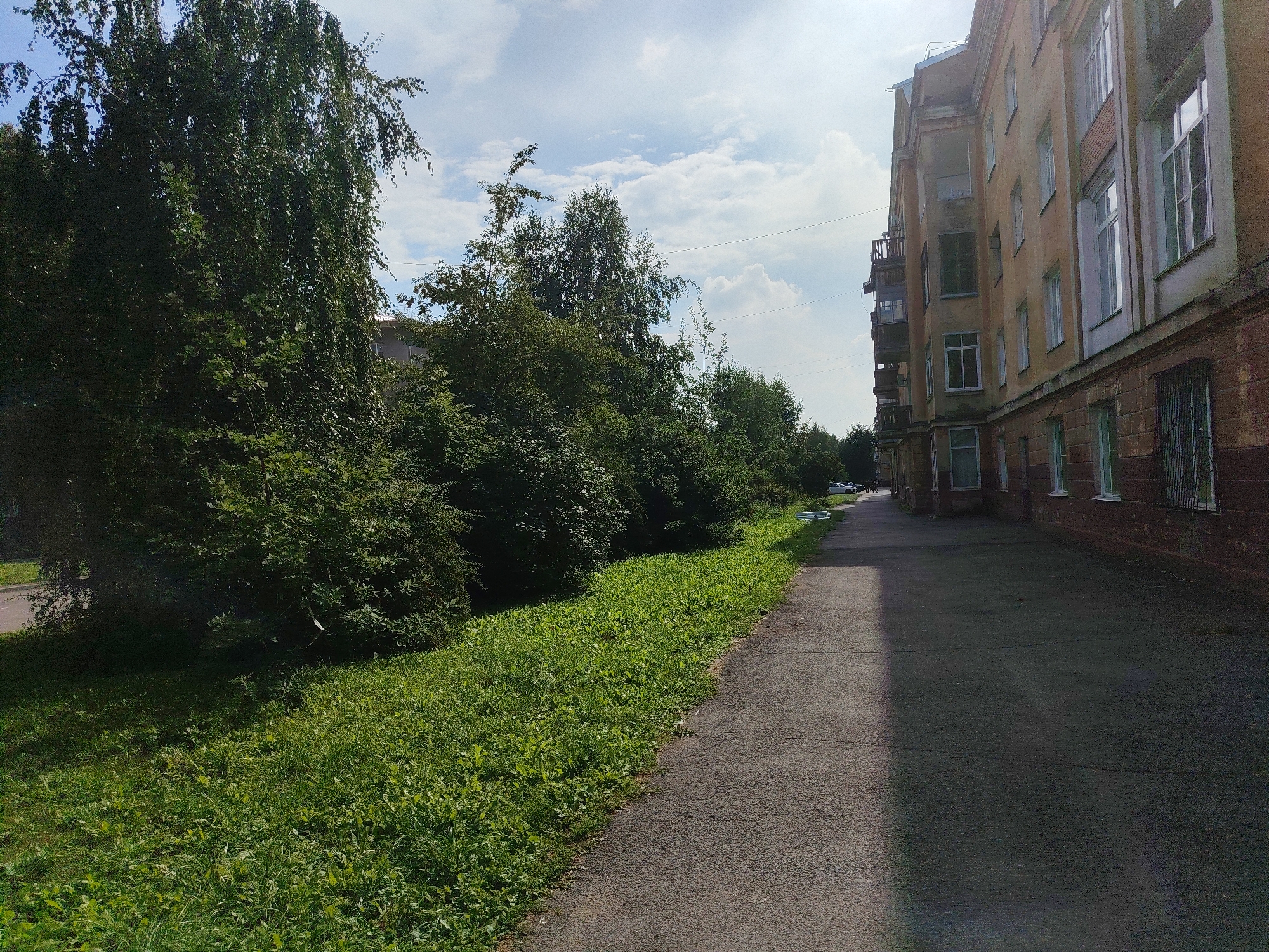 The image shows a paved walkway running alongside a grassy area and a multi-story building. The building appears to be made of brick and has multiple windows and balconies. There are trees and foliage along the left side of the walkway. The sky is partly cloudy. The scene is likely a residential area.