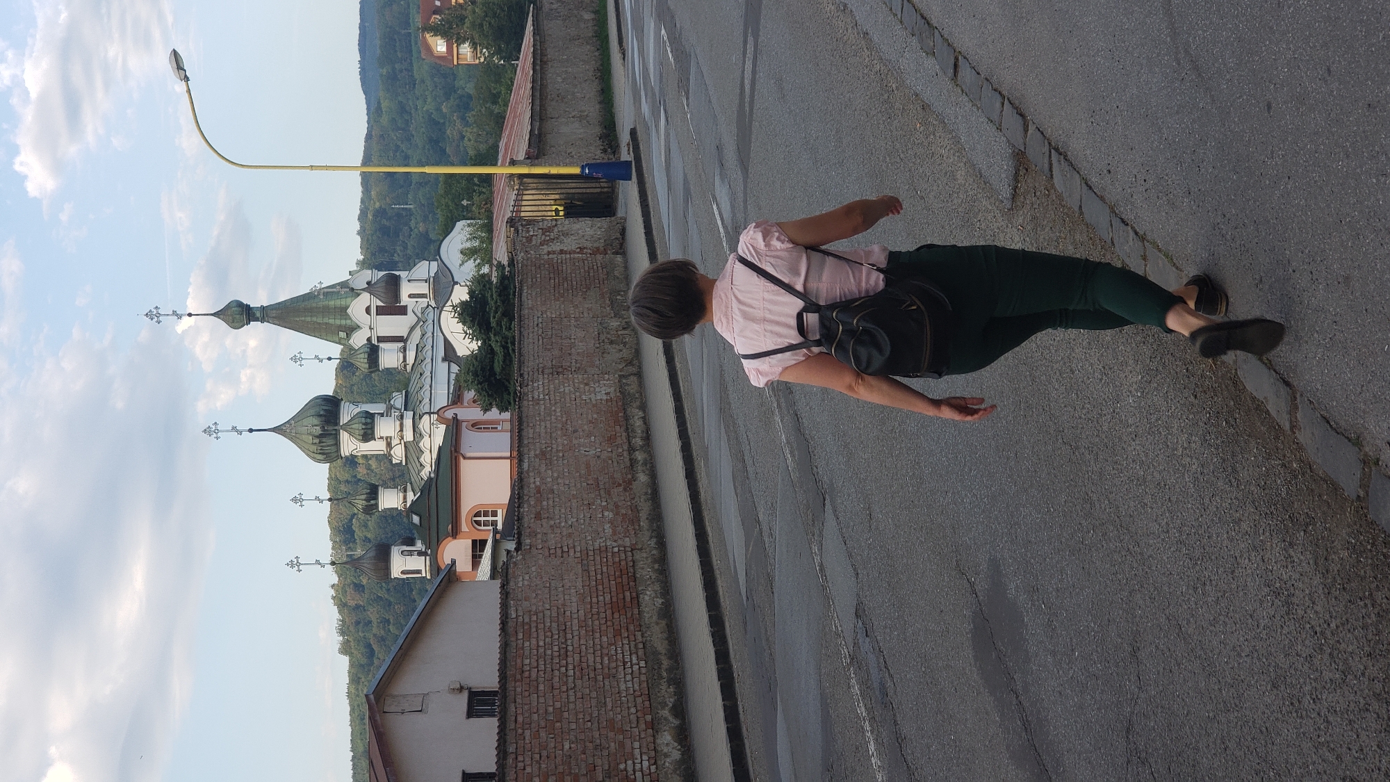A woman with short, dark hair walks away from the camera down a paved road in Prešov, Slovakia. She is wearing a light pink, short-sleeved collared shirt, dark green pants, and black flats. A black backpack is slung over her shoulders. The road has visible cracks and patches. A sidewalk with a stone curb runs along the right side of the road. In the background, a brick wall encloses a property with buildings and trees. Dominating the background is a large, white and green domed church with multiple spires, characteristic of Orthodox architecture. A yellow lamppost with a modern light fixture stands tall on the right side of the frame. The sky is partly cloudy, with a light blue hue. The scene appears to be set during the daytime, possibly late afternoon.