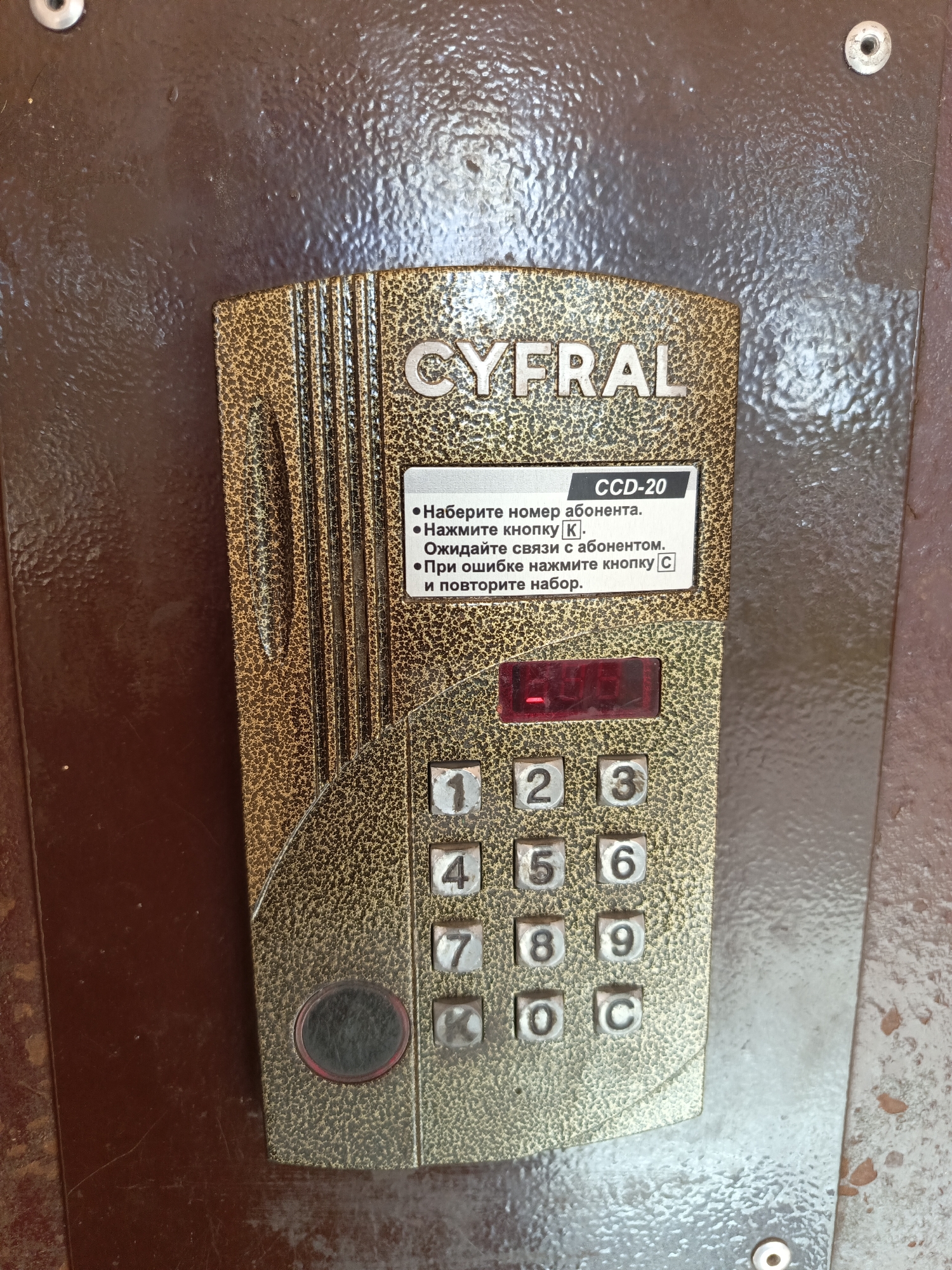 A close-up, eye-level, exterior shot captures a detail of a gold-colored, textured intercom panel with a dark brown, weathered, textured metal wall as the background. The intercom panel, which appears to be made of metal, has a patterned surface and features the brand name "CYFRAL" embossed at the top. Below the brand name, a white rectangular label displays "CCD-20" and instructions in Russian, indicating: "Dial the subscriber's number. Press the [K] button. Wait for the subscriber to connect. If there is a mistake, press the [C] button and redial." A red digital display panel is situated below the label, with no visible numbers illuminated. A standard numeric keypad with buttons numbered 1 through 9, plus 0, K, and C, is arranged in a grid. A circular button or speaker, possibly metallic with a red rim, is located at the bottom left of the keypad. The wall behind the intercom is a dull, dark brown with a rough, uneven texture and shows signs of wear and possible moisture. Two small metal screw heads are visible on the wall, one at the top left and another at the bottom right. The overall scene suggests an outdoor, urban environment, possibly an entrance to a residential building. The location is Ivatsevichy, Belarus, a small city in Brest Region.