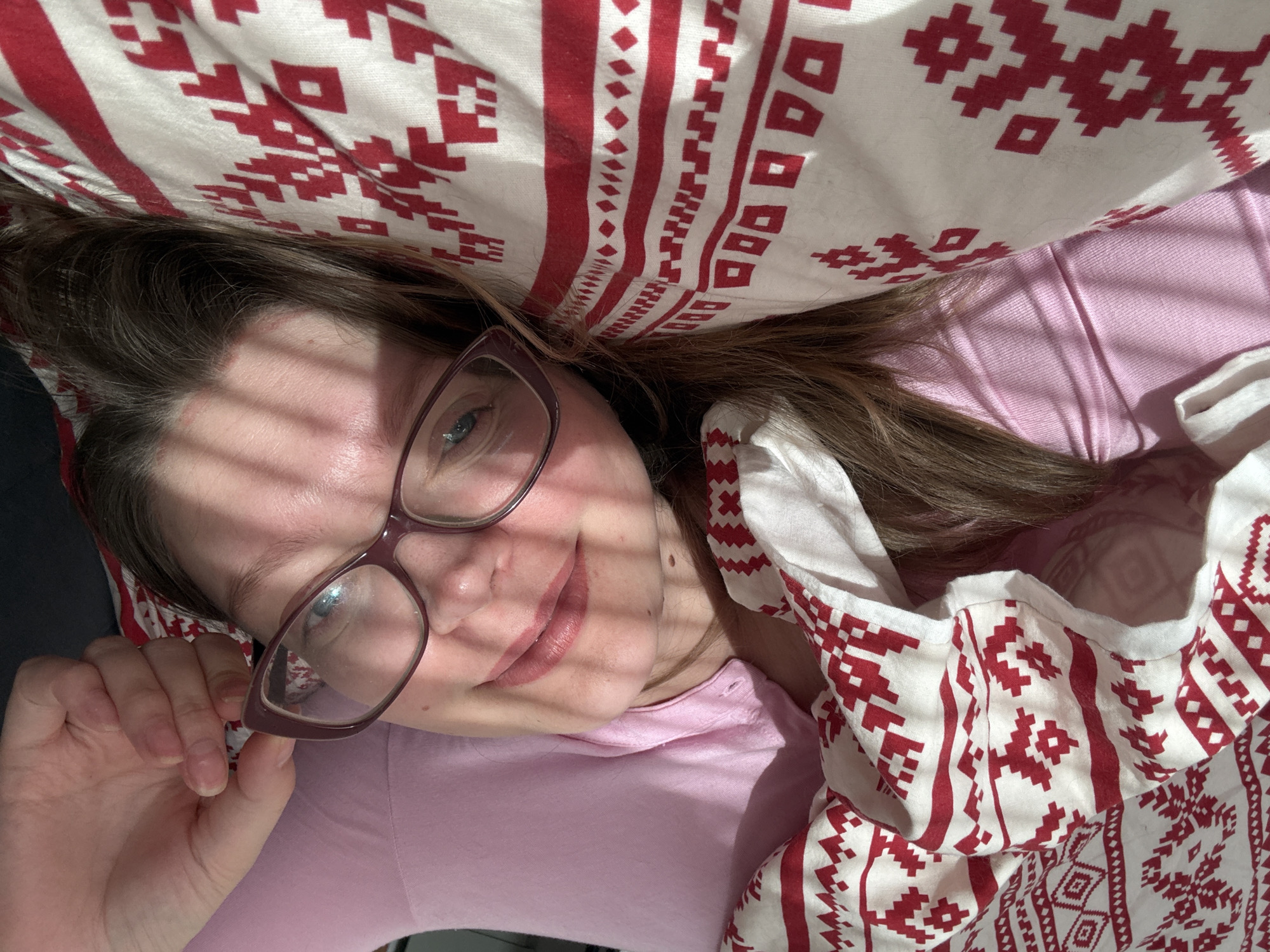 A woman with brown hair and glasses lies on a bed. The bed has a patterned red and white pillow and blanket. The woman is wearing a pink shirt. The image is taken from an overhead perspective and shows the woman's face and upper body. Light shines across her face, creating stripes of light and shadow. The location is unknown.