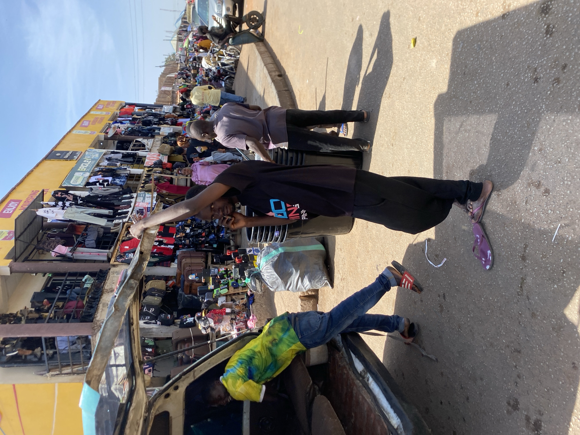 The image depicts a bustling outdoor market scene in Bukuru, Nigeria. People are present along a street lined with stalls selling clothing and other goods. A man in a black shirt is standing and talking on a phone. Another man in a purple shirt is standing nearby. A person wearing a yellow and blue shirt is seen reaching inside a car. A large gray sack is on the ground. Other objects visible include bags, luggage, and various items for sale at the market stalls.