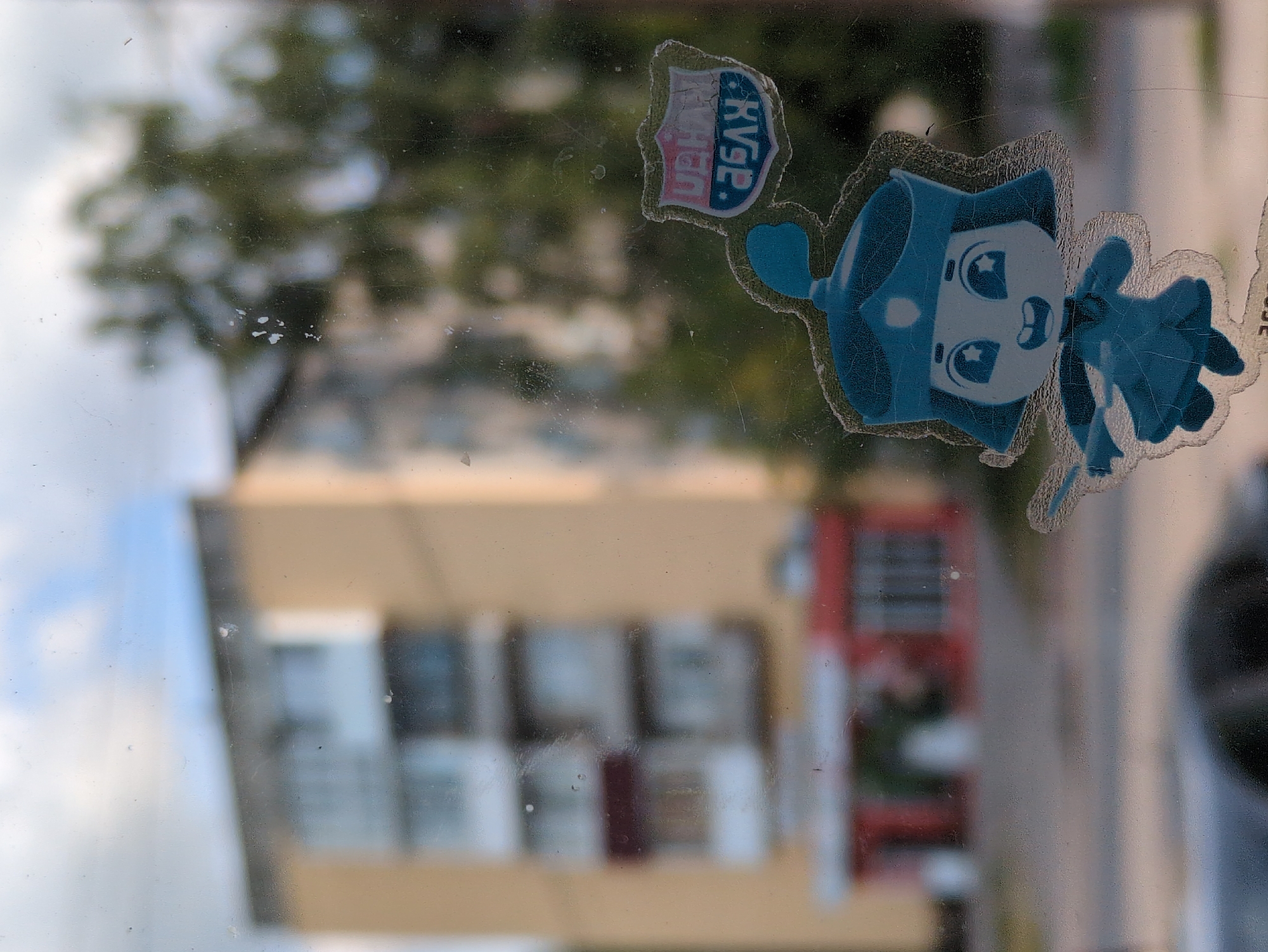 This is a photograph taken through a dirty window in Kulebaki, Russia. The window has a sticker of a cartoon character in the lower right corner. The character is blue, with a crown, large eyes with star pupils, and an open mouth. The character is holding a staff with a blue flame at the end. Behind the sticker, through the window, a blurry yellow apartment building with balconies is visible. To the right of the apartment building, a blurry green tree is seen. In the foreground, a small shield-shaped sticker with Russian text, mostly illegible due to blur and angle, is also present. The environment suggests an urban setting with residential buildings and foliage.