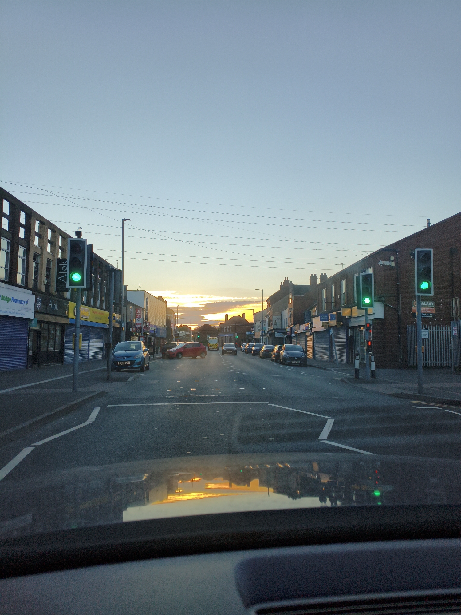 The image shows a view from a vehicle driving down a street at dusk. The street is lined with shops and buildings on both sides, with traffic lights visible. The sky is clear with some clouds on the horizon, and the setting sun casts a warm glow. The road surface is asphalt, with white markings. The reflection of the sky and buildings can be seen on the vehicle's dashboard. This scene is located on Walsall Road in Sandwell, United Kingdom. There are no people visible in the image.