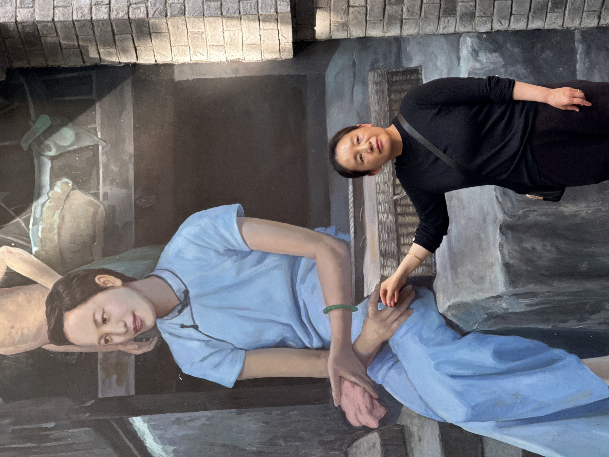 A real woman poses playfully with a large, realistic mural depicting a traditional scene, likely a form of 3D street art or trompe l'oeil. The image is oriented vertically.
The real woman, of East Asian descent, stands to the right of the mural, looking towards the camera with a soft, neutral expression. She wears a long-sleeved black top and black trousers, with a dark strap draped over her right shoulder. A gold ring is visible on her right hand. She interacts with the painted figures; her right hand gently rests on the knee of the main painted woman, and her left hand is positioned above the painted woman's upper arm, near what appears to be a wicker basket texture.
The mural itself portrays a tranquil, older-style setting. The prominent painted figure is a young East Asian woman, depicted in a relaxed, almost reclining pose. She wears a light blue traditional top with dark fastenings and a light blue skirt or trousers. A green jade bangle adorns her left wrist. Her head is gently rested on her right hand, and she smiles serenely. Her left hand is placed on the foot of another painted figure, only partially visible as a bare torso lying on its back to the left.
The painted background features dark, shadowed areas, with some lighter details suggesting architectural elements like walls or beams, and brickwork visible along the top edge of the mural. The overall setting implies a historical or cultural context typical of Nan'an District, China, where such art installations often reflect local heritage. No visible text is present in the image. The scene appears to be illuminated by natural daylight.