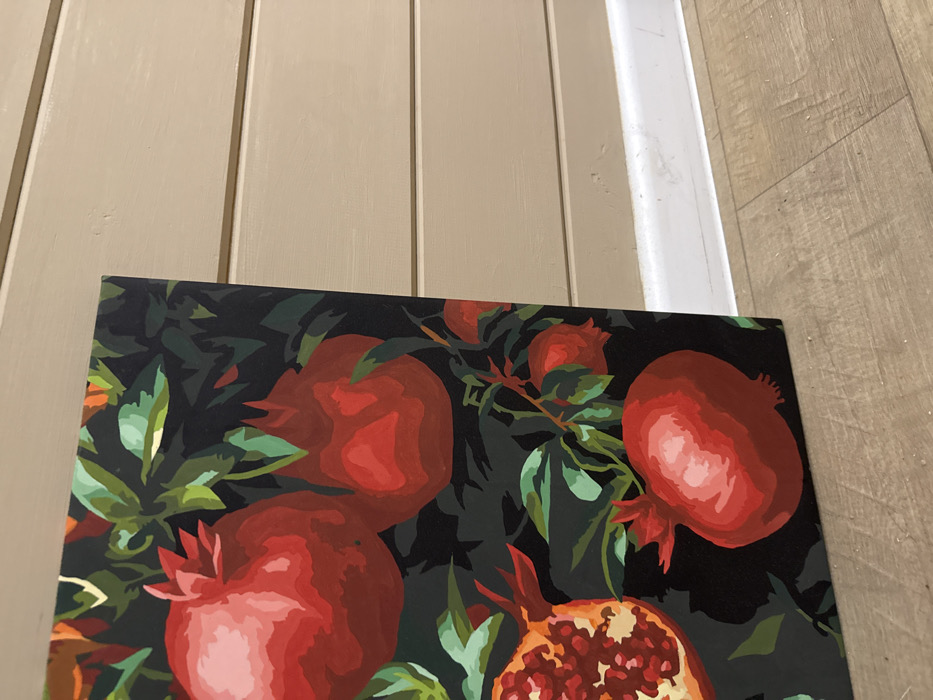 An indoor scene is depicted, featuring a vertically oriented canvas painting leaning against a paneled wall. The painting occupies the left portion of the frame and portrays a stylized arrangement of red pomegranates and green leaves on a dark background. Multiple whole pomegranates are visible, along with one partially cut pomegranate revealing its red seeds. The wall, located to the right of the painting, is light beige or pale taupe, constructed from horizontal planks. A white baseboard runs along the bottom of the wall, meeting a light brown, wood-patterned floor. No individuals or discernible actions are present. The specific location within the city of Chaikovsky, Russia, cannot be determined from the visual evidence.