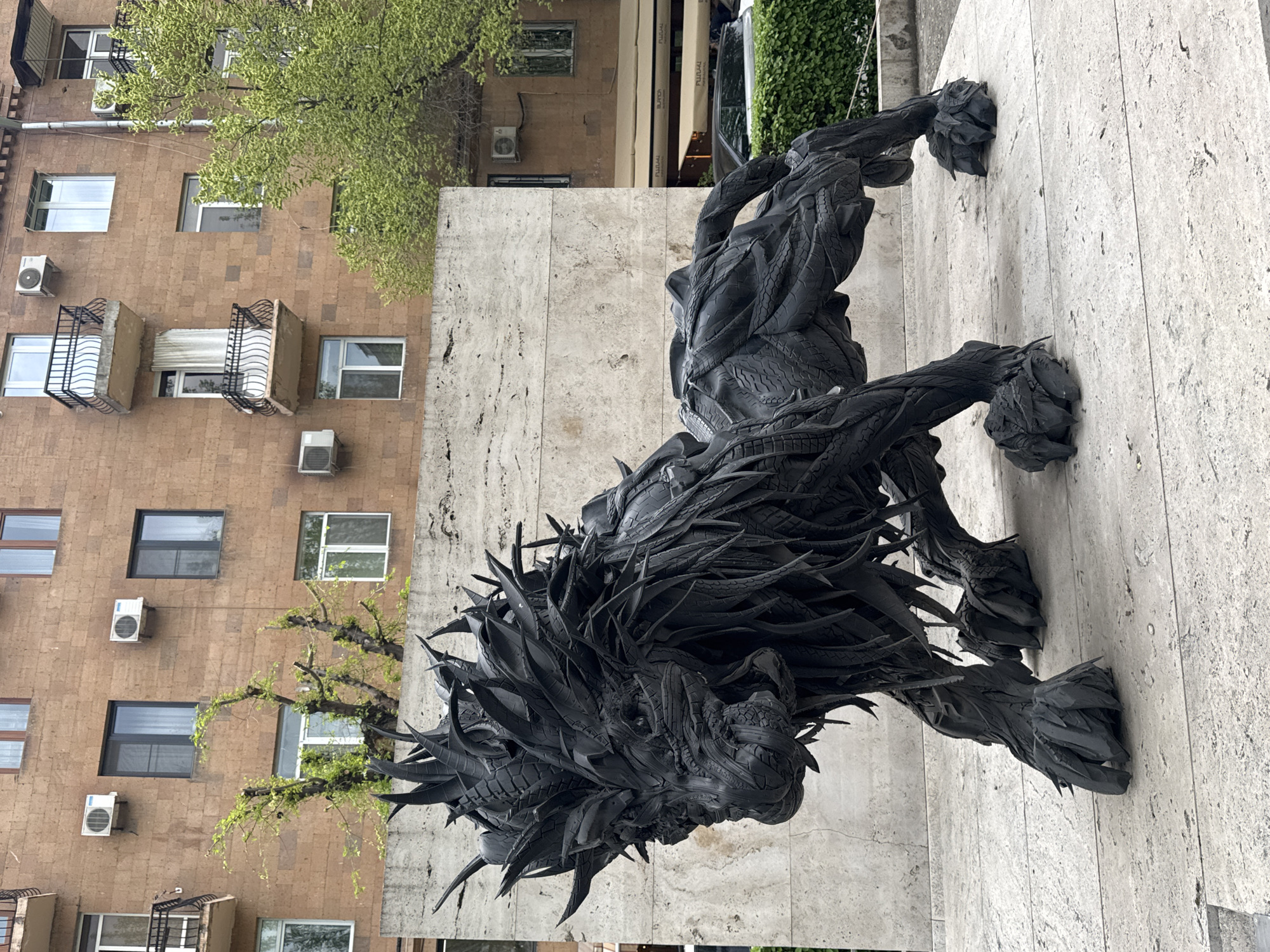 The image features a large, dynamic sculpture of a lion-like or mythical creature, crafted predominantly from recycled dark rubber, resembling tire treads and cut rubber pieces. The beast is depicted in a powerful, walking or stalking pose, with a heavily textured body and a dramatic, spiky mane. The sculpture stands on a pale, light-colored stone or concrete wall that serves as its base, suggesting a public art installation.
Behind the sculpture, a multi-story urban building with a brown, tiled facade dominates the left side of the frame. Numerous windows, some reflecting the bright sky, and several white external air conditioning units are affixed to its exterior. Two small balconies with decorative black railings are also visible. A vibrant green tree with delicate, light-green leaves partially overlays the building, adding a touch of nature to the urban setting. In the distant background, subtle hints of other buildings, greenery, and possibly a street are discernible, including text that appears to read 'PULMAN' on a commercial awning. The scene is illuminated by natural daylight, suggesting mid-day. The photograph captures an interplay between repurposed materials, urban architecture, and nature in Yerevan, Armenia.