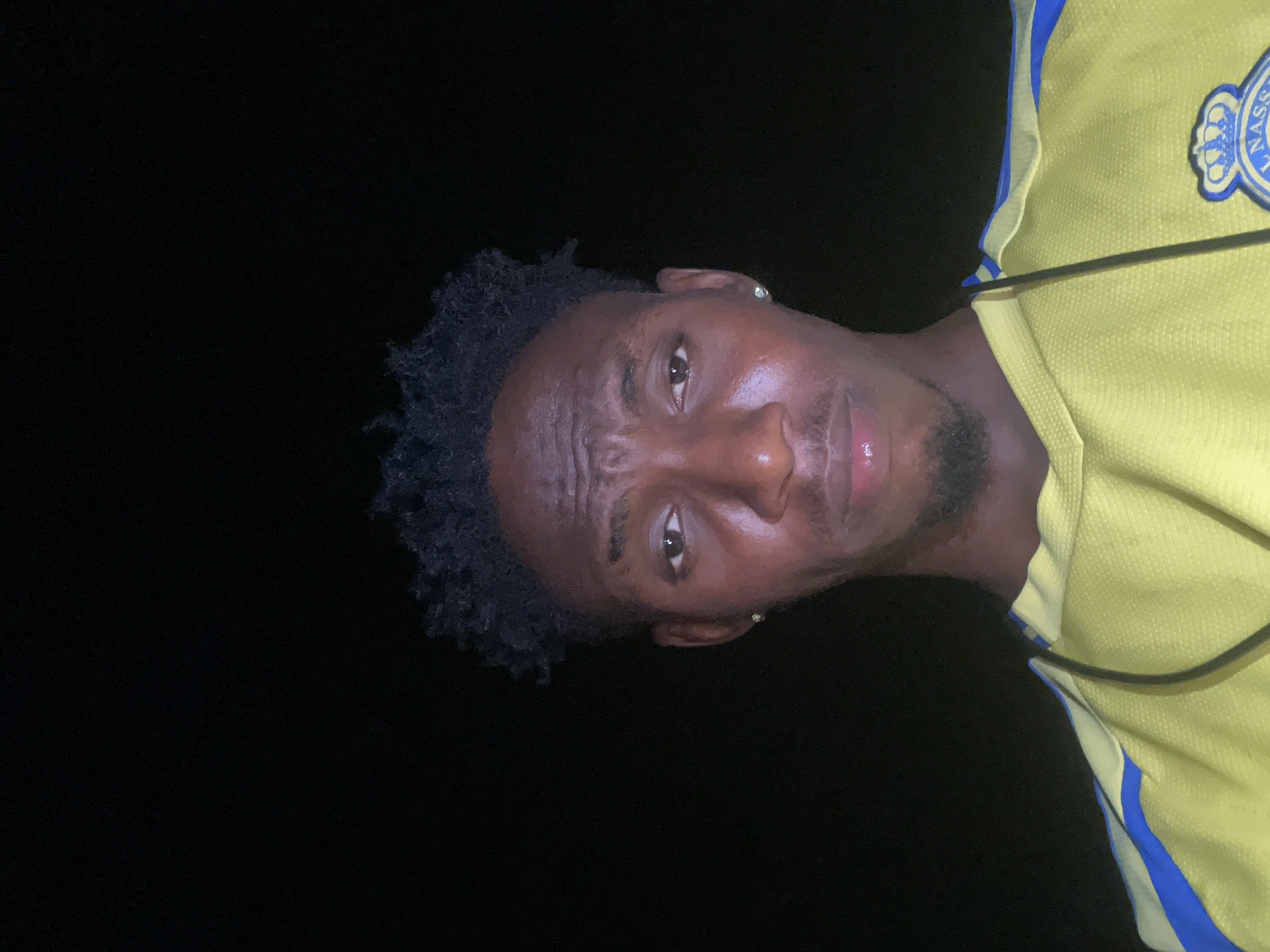 A man with dark skin is pictured against a black background. He is wearing a yellow shirt with blue trim and a logo on the upper right side. The lighting suggests it is dark, possibly nighttime. The location, Lokoja, Nigeria, cannot be confirmed from the image alone.