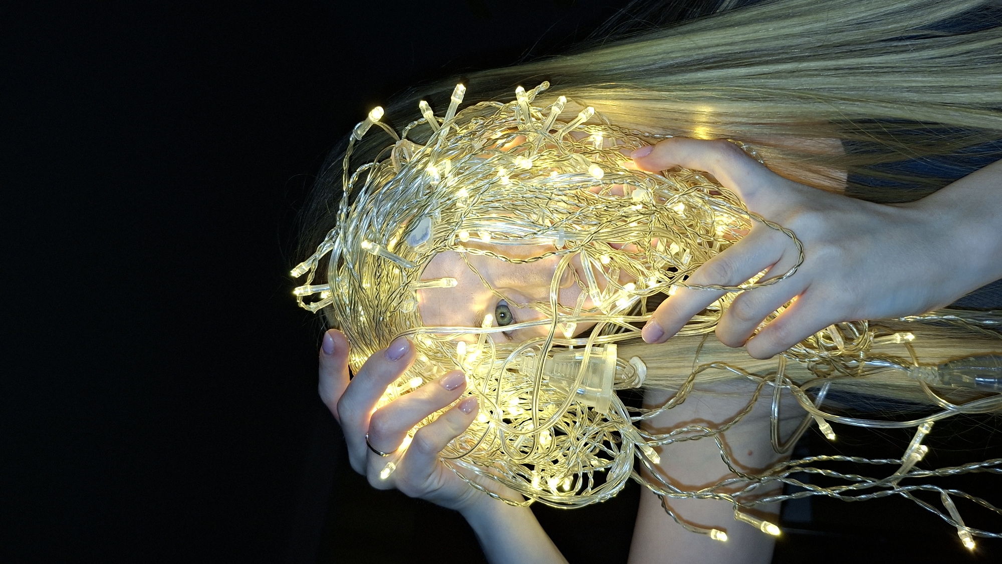A woman with light blonde hair is holding a large tangle of warm white fairy lights in front of her face. Her eyes are visible through the lights. The background is dark. The location is not discernible from the provided image.