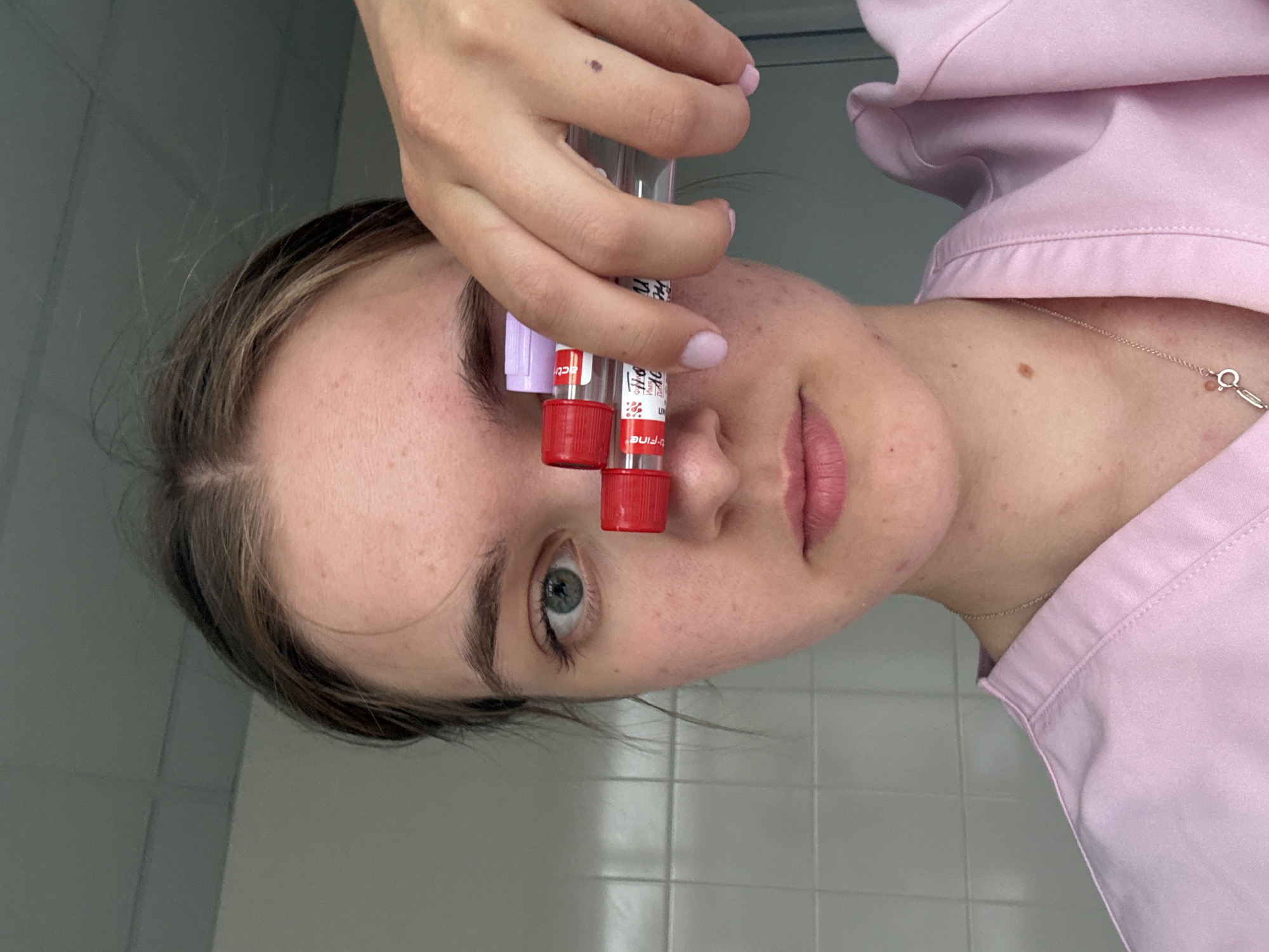 A fair-skinned woman in a light pink collared shirt is holding two blood collection tubes with red caps in front of her face. The tubes obscure her nose and part of her eye. The woman's eye is a light blue-green color. She has light brown hair with blonde highlights, and is wearing a delicate necklace. The background appears to be a tiled wall, potentially in a medical or laboratory setting. There is no explicit indication this picture was taken in Perm, Russia.